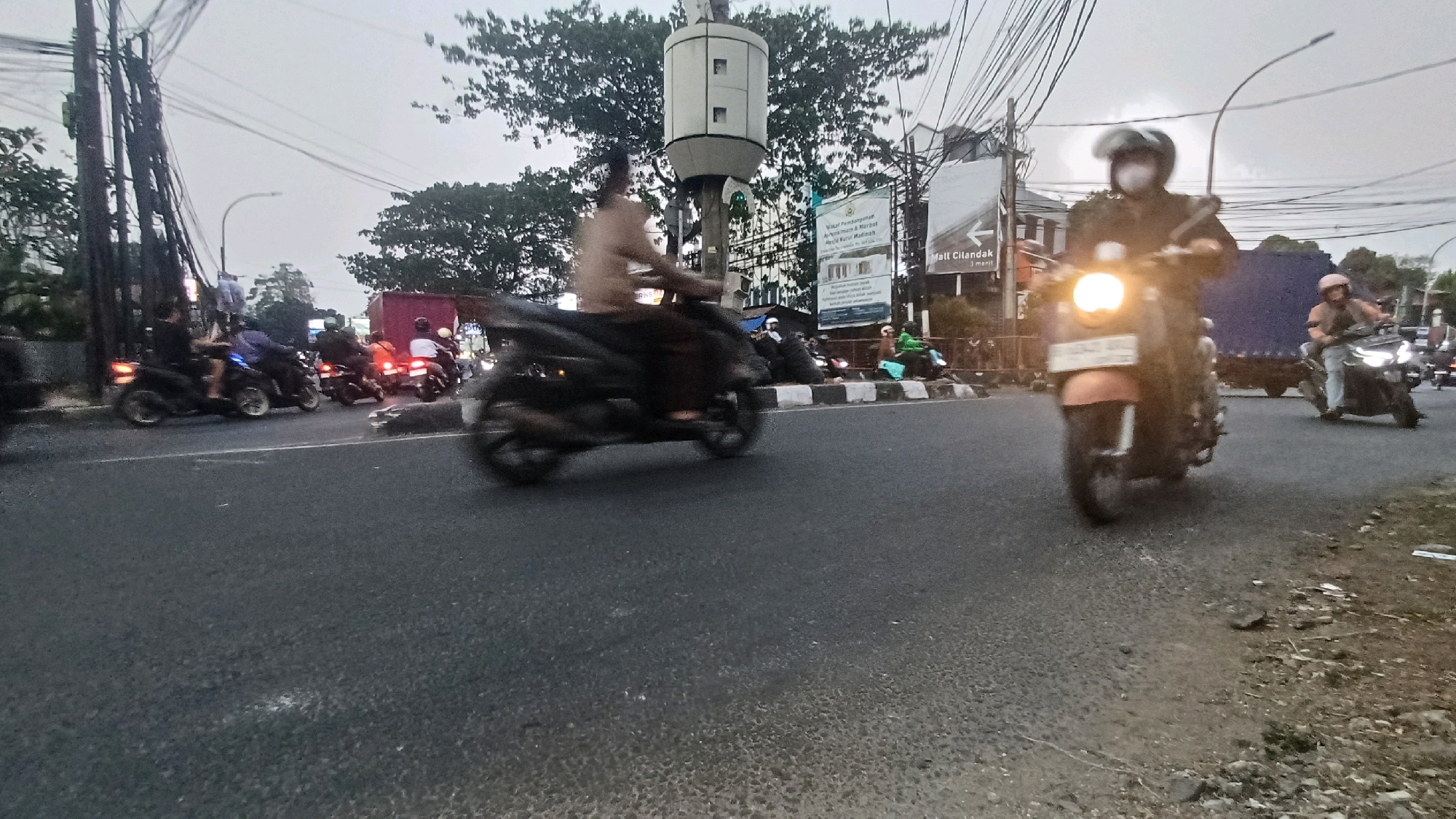 The image depicts a busy street scene in South Jakarta, Indonesia, likely during overcast conditions or twilight. The foreground features an asphalt road with several motorcycles in motion, indicated by motion blur. A prominent orange scooter with its headlight illuminated is moving towards the viewer. To the right, another motorcycle with a rider wearing a helmet is also moving away. In the midground, a black motorcycle with a blurred rider is crossing the frame from left to right. Further back, other motorcycles and a red truck are visible, suggesting traffic congestion. The background is characterized by a dense network of electrical wires overhead, trees, and buildings, including a billboard with Indonesian text. A tall, cylindrical structure with white panels, possibly a traffic monitoring device or utility pole, stands near the center. The overall impression is a snapshot of daily urban life in a bustling Indonesian city.