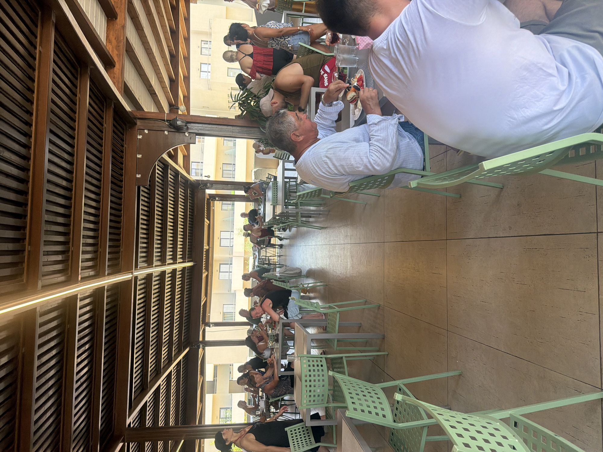 An outdoor dining area at a resort in Georgioupoli, Greece, is captured in this medium-angle shot. The scene is populated with patrons enjoying a meal under a wooden-slatted roof structure. In the foreground, a man wearing a white shirt is actively eating a burger, while another man in a white t-shirt is seated to his right, his back to the camera. Several light green chairs are strategically placed around tables. In the mid-ground, a group of individuals are seated at tables, engaged in conversation and dining. The background reveals a building with balconies, characteristic of a resort. The overall ambiance suggests a relaxed atmosphere typical of a vacation setting.