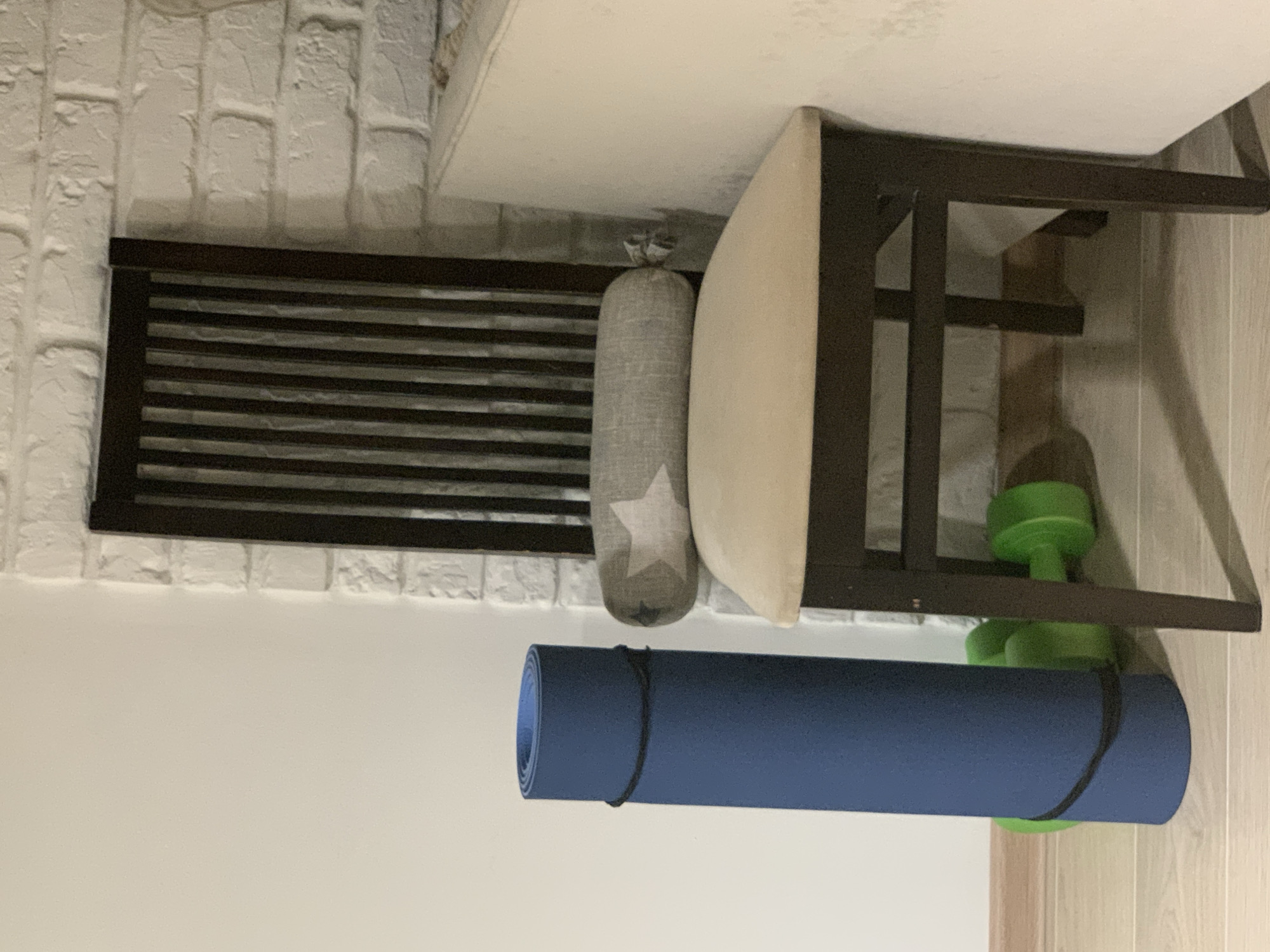 In Kapyĺ, Belarus, a dark wooden chair with a beige cushioned seat and a tall, slatted back stands against a white brick wall. A grey bolster pillow with a white star pattern rests on the chair's seat. To the left of the chair, a rolled-up blue yoga mat is propped upright and secured with black bands. Two bright green dumbbells lie on the light-colored wooden floor in front of the yoga mat. The scene suggests a home environment dedicated to fitness.