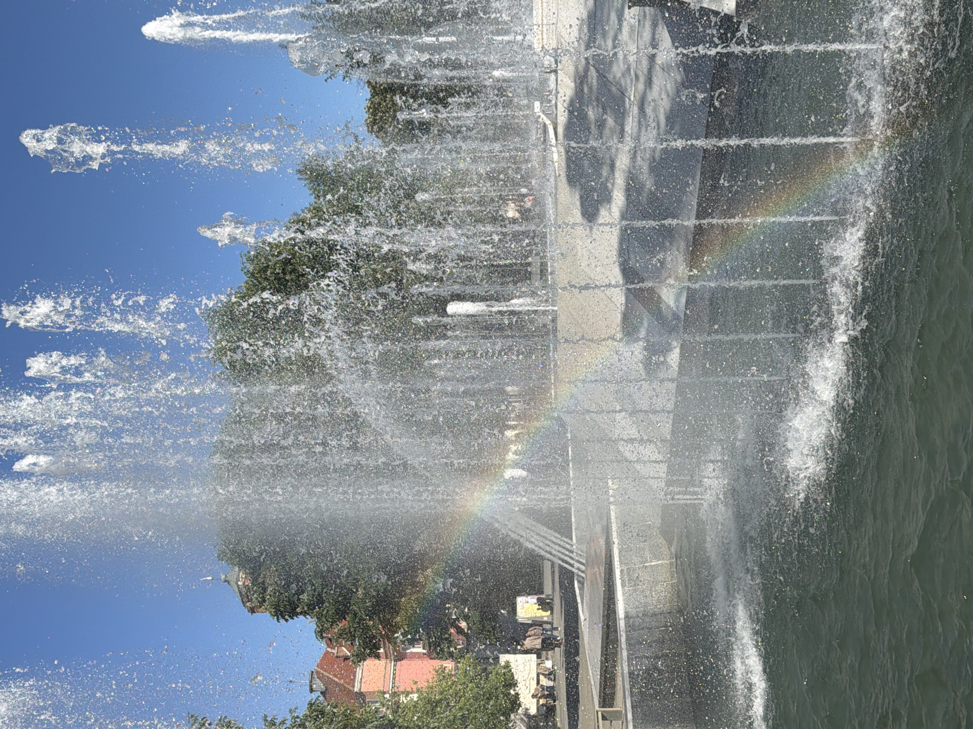 A full shot of a water fountain with a partial rainbow visible within the spray. The fountain is gray and tiered, with water cascading over the sides. A body of water is directly to the right of the fountain. Trees and a building with a red roof are visible through the fountain spray. The sky is blue. The location is Pleven, Bulgaria.