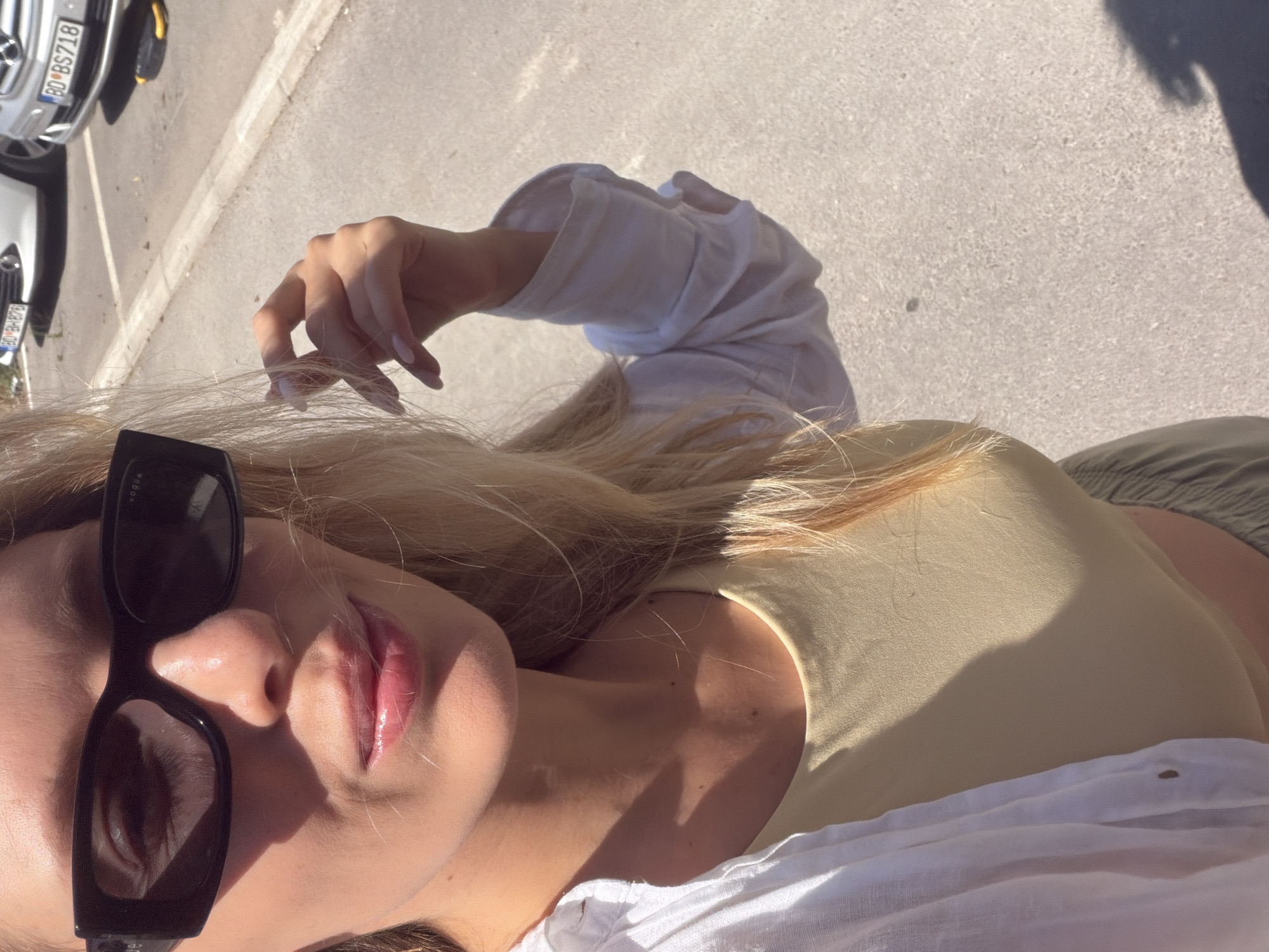 A young blonde woman with fair skin is captured in a close-up, outdoor selfie, holding her right hand up towards her face. She is wearing large, black, rectangular sunglasses with the "Vogue" logo visible on the temple. Her lips are glossy and slightly parted. She has on a light yellow, sleeveless top and a loosely buttoned, white open-collared shirt over it. Her blonde hair falls loosely around her shoulders. The background shows a paved area, likely a parking lot, with several cars parked. The license plates of two visible cars read "BD-BH876" and "BD-BS718". The scene is illuminated by bright sunlight, casting shadows on the ground and the woman's face. This image was taken in Budva, Montenegro.