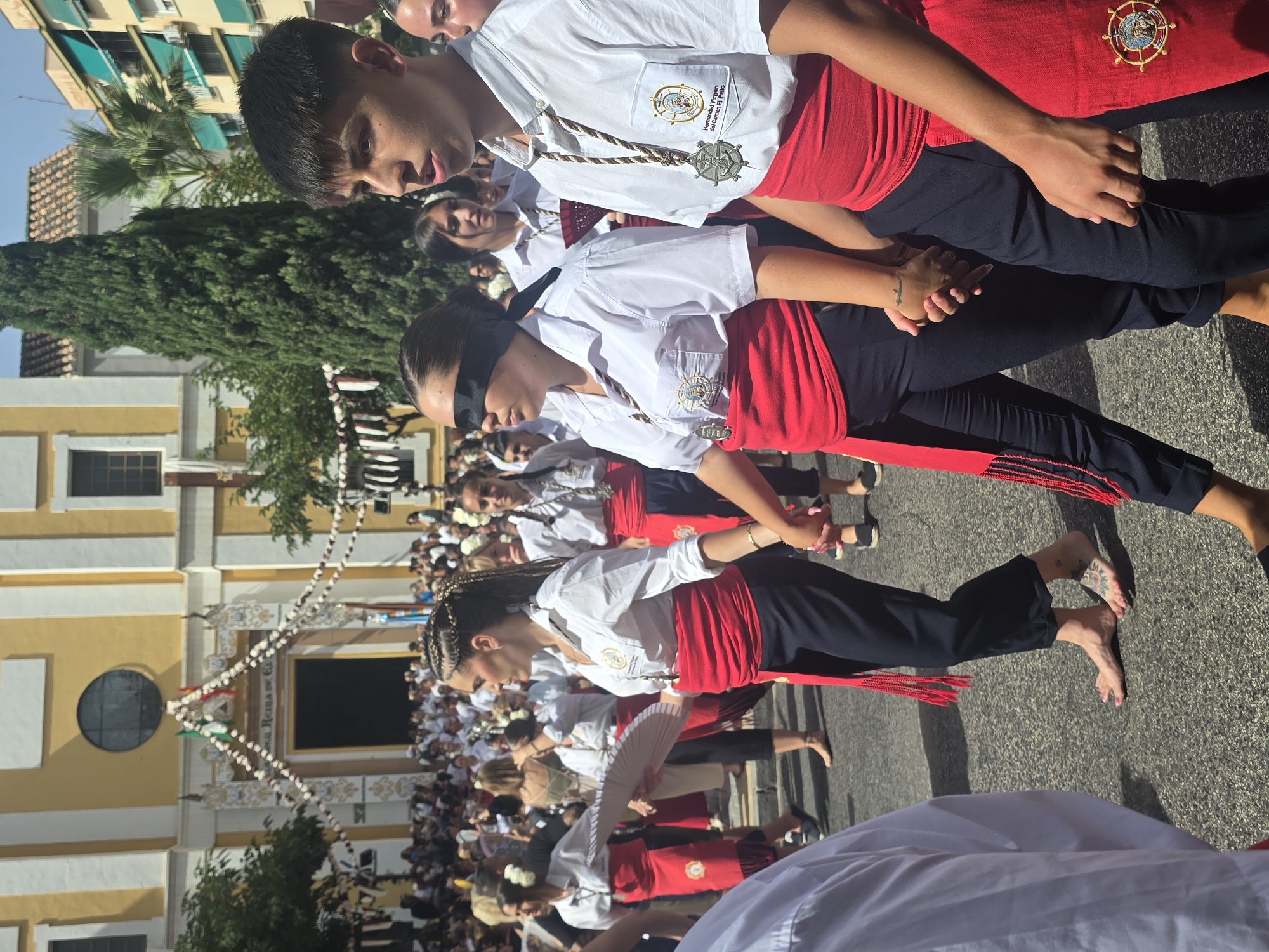 In a sun-drenched street in Málaga, Spain, a procession is underway in front of a yellow and white building adorned with festive garlands. The central figures are young people, predominantly women, dressed in white shirts and dark trousers, cinched with wide, vibrant red sashes. Several of the women have their hair styled in intricate braids, and some wear white flowers in their hair. One woman in the foreground is blindfolded with a black cloth, her hands held by another person. Another woman is carrying a large, white fan. Both the blindfolded woman and the person next to her are wearing large circular medallions around their necks. The procession appears to be moving forward, with a crowd of onlookers visible in the background. The building behind them has a large arched entrance, suggesting it might be a church or public building. The overall atmosphere suggests a cultural or religious event.
