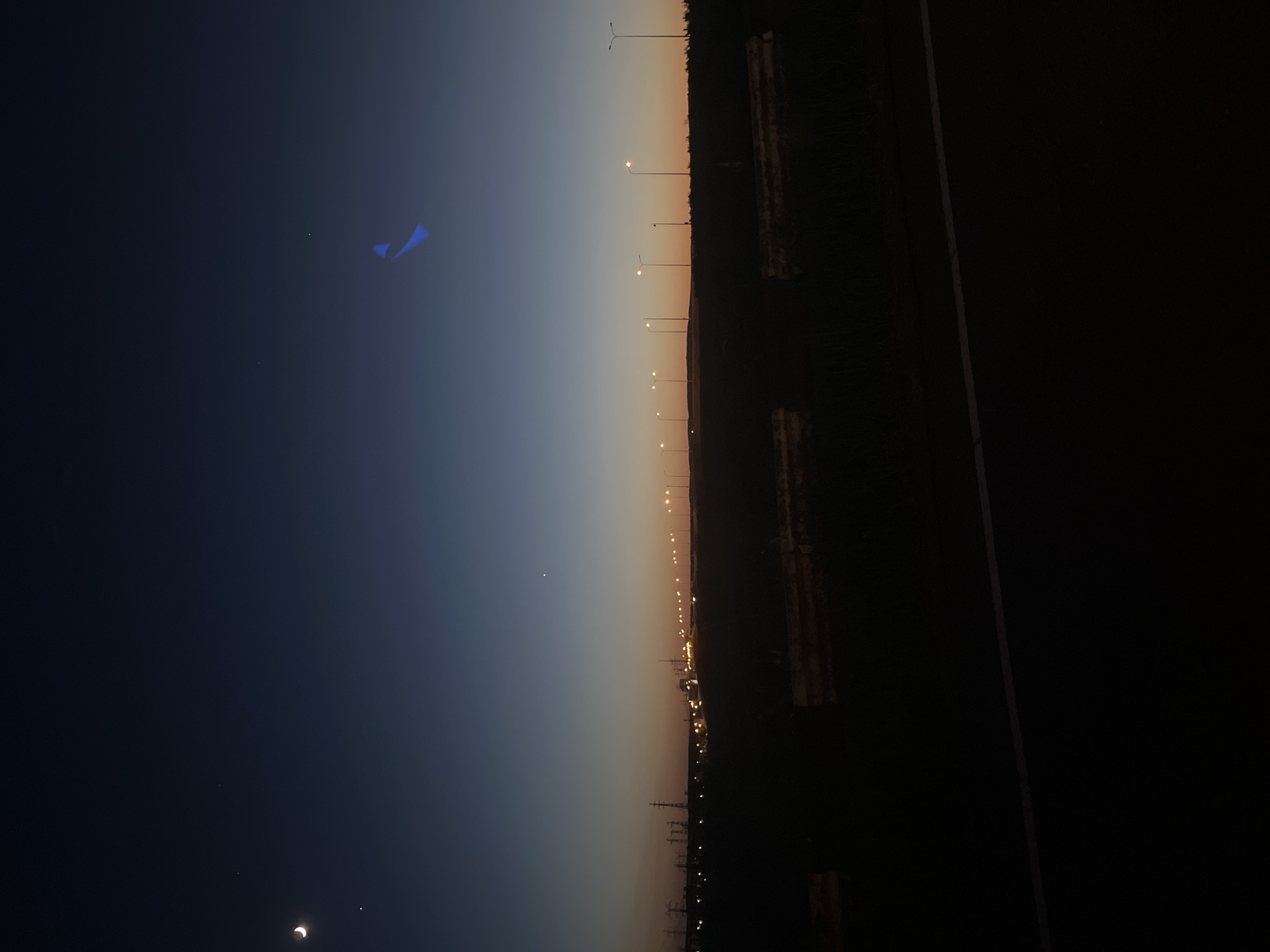 The image depicts a view at dusk or dawn in or near Chaikovsky, Russia. The horizon fades from dark blue to a lighter orange/yellow. A road or path runs along the right side of the frame, in shadow, with trees or vegetation visible between street lights running along the side of the road. In the dark sky, there are blue streaks and some stars, as well as what appears to be a partial eclipse of the moon. It is devoid of any identifiable people or actions. The environment consists of a city or town adjacent to a road or path at a low light level.