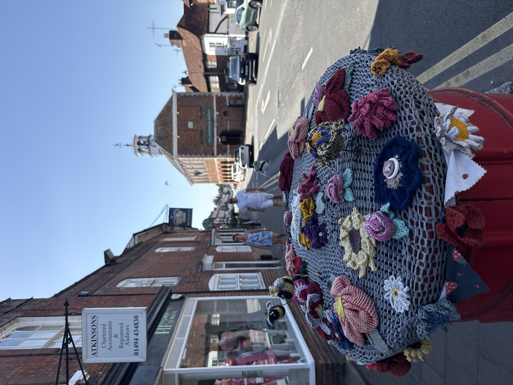 The image depicts a street in Amersham, United Kingdom. A bollard covered in a knitted grey net adorned with various colorful crocheted flowers is in the foreground. In the background, on the left side of the street, are brick buildings with storefronts. A sign for "Atkinsons Chartered Accountants & Registered Auditors" is visible. People are walking on the sidewalk in the distance. Across the street, a brick building with a clock tower is visible. Cars are parked on the street. The sky is blue.