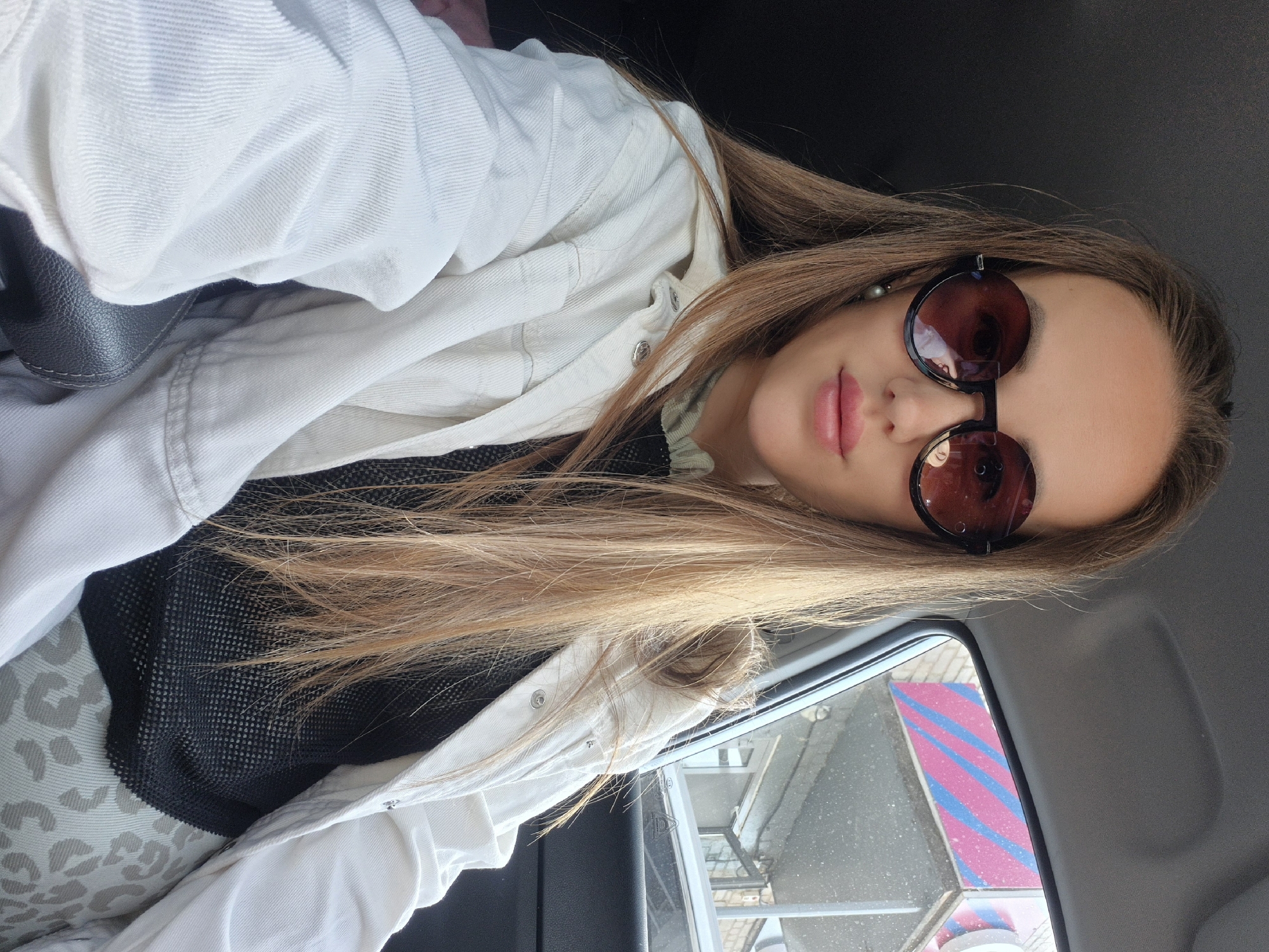A selfie shows a young woman in a car in Unknown, Unknown. She has long, light brown hair and is wearing round, brown sunglasses. She has pink lipstick. She is wearing a white jacket over a black mesh top and a grey and white patterned garment. The interior of the car is visible, as is the side window, showing a building with pink and blue stripes.