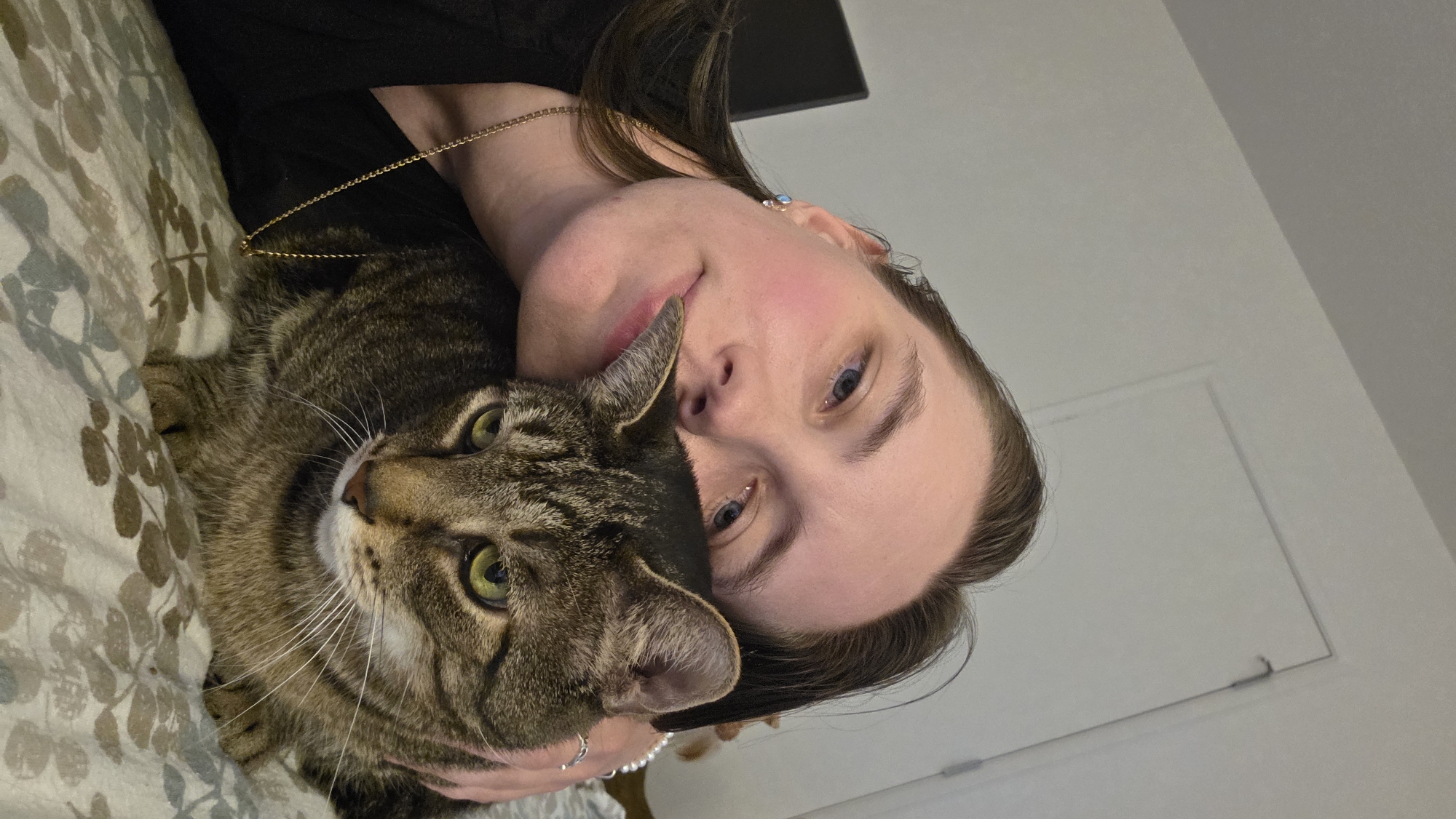 A selfie shows a person with light skin and blue eyes, with brown hair, resting their head on a tabby cat. The person is wearing a black shirt and a gold necklace. The cat has green eyes and is looking towards the camera. The background consists of a white wall and a door. The setting appears to be indoors, likely a bedroom. There is no visible indication of the specific location within Portland, United States of America.