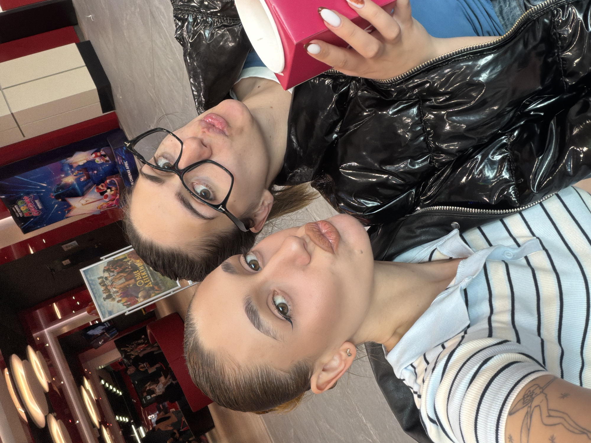 Two young women are posing for a selfie in what appears to be a cinema or entertainment venue. The woman on the left wears glasses and a black shiny jacket, holding a pink object in her hand, and is positioned slightly behind the other woman. The woman on the right has her hair pulled back and wears a striped top. The background includes posters and illuminated circular designs on the walls, suggesting an entertainment or public space. Given the presence of film posters in the background, the location might be a cinema hall or lobby. There are no indicators suggesting the city of Sofia, Bulgaria.