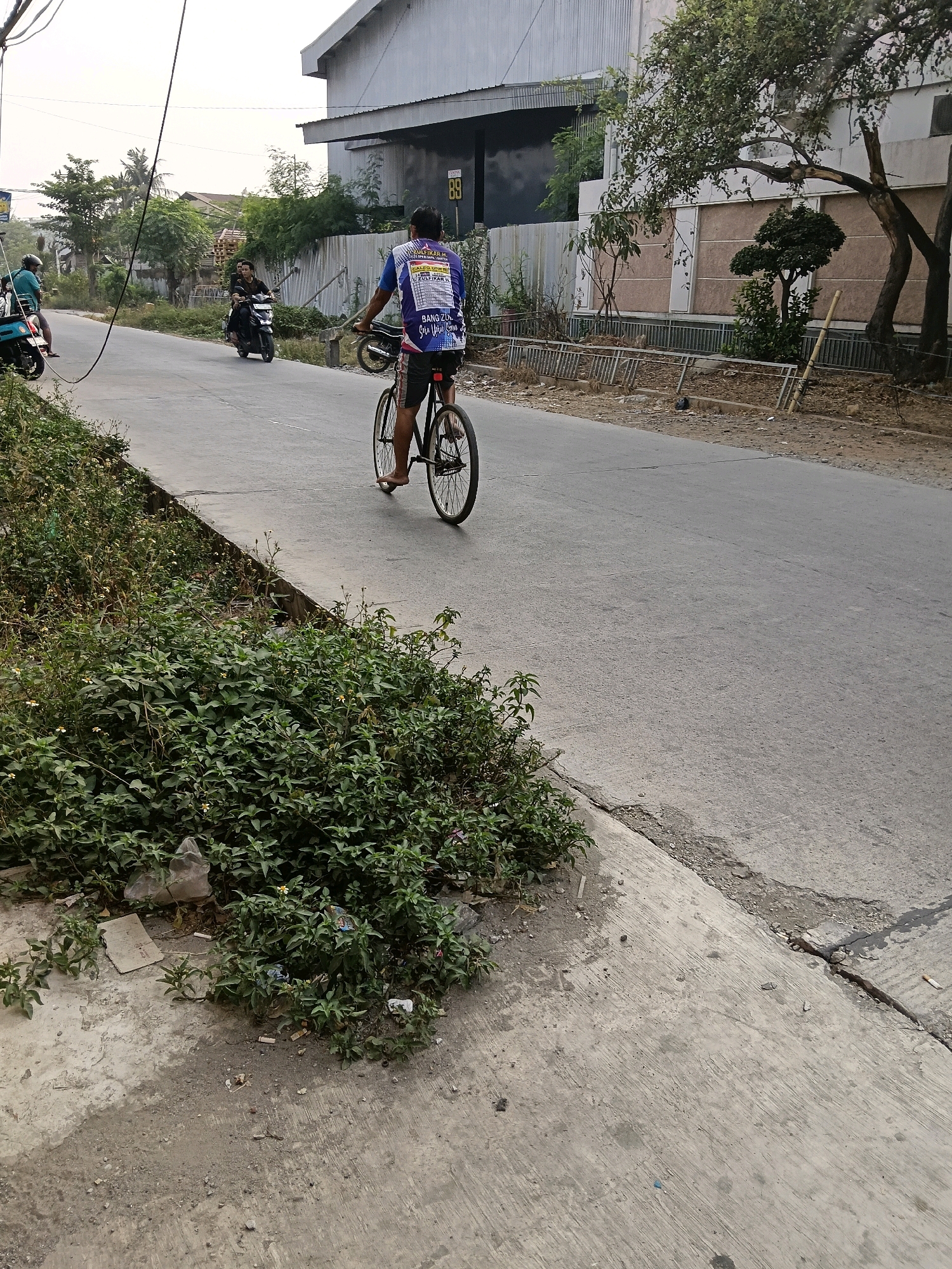 In Belimbing, Indonesia, a man on a bicycle rides on the right side of a paved road, facing away from the camera. He wears a blue and white t-shirt with text and dark shorts, and is barefoot. Behind him, a motorcycle carries two passengers, and further in the background, another motorcycle is visible with one rider. The road is bordered on the left by a concrete walkway and dense green vegetation with scattered white flowers and debris, including plastic bags and cigarette butts. On the right side of the road, a large building with a corrugated metal exterior and a lighter-colored front section stands, partially obscured by trees. A fence runs along the front of the property. The sky is overcast.