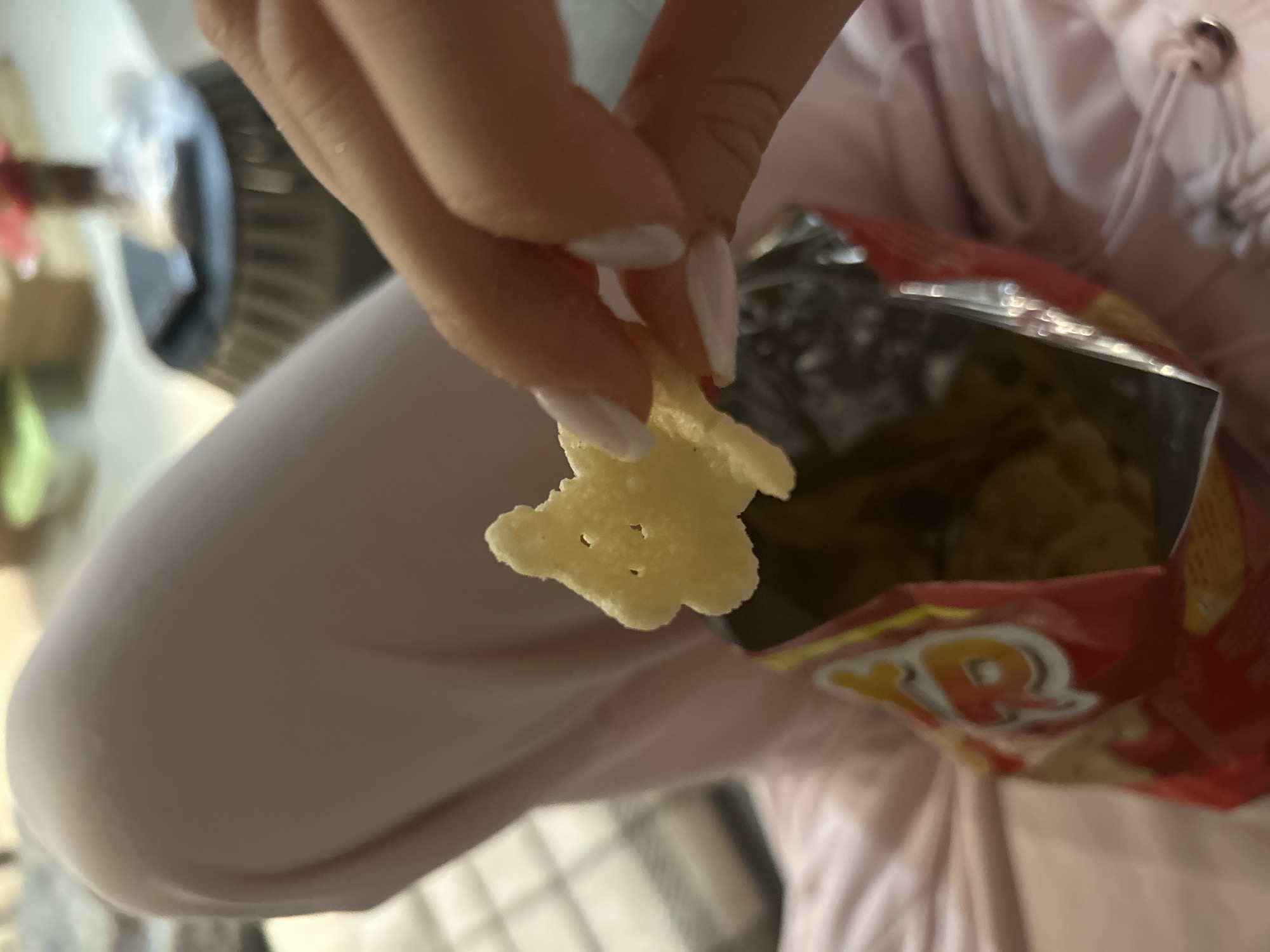 A close-up shot depicts a person's hand holding a yellow chip in front of an open bag of chips. The person's hand has long white nails. The bag of chips is red and yellow with the brand name "R" visible. The background includes a light pink garment and a blurred view of what appears to be a chair and other objects. The location is unknown.