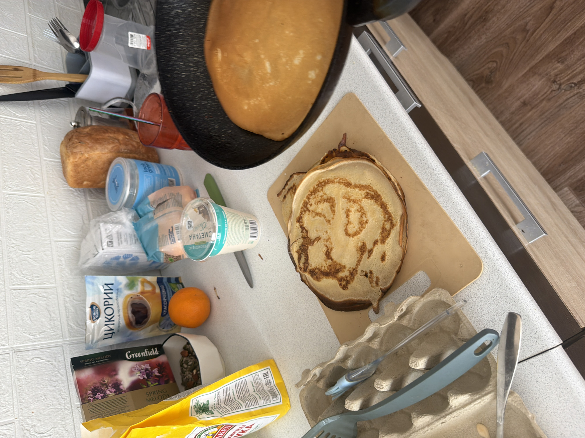 The image depicts a countertop scene in what appears to be a kitchen setting. Food items, containers, and utensils are scattered across the surface. There's a frying pan containing a pancake or crepe, next to a stack of pancakes on a cutting board. Other visible items include a loaf of bread, a container of "СМЕТАНА" (sour cream), a bag of "ЦИКОРИЙ" (chicory), tea boxes, an orange, and a package of "Мука" (flour). Kitchen utensils such as a spatula, fork, and knife are also present. An egg carton with cooking utensils is near the pancakes on the cutting board.
There is no information in the image to indicate a location in the city of Leningradskaya, Russia. The presence of Russian products does not explicitly place the image in that city.