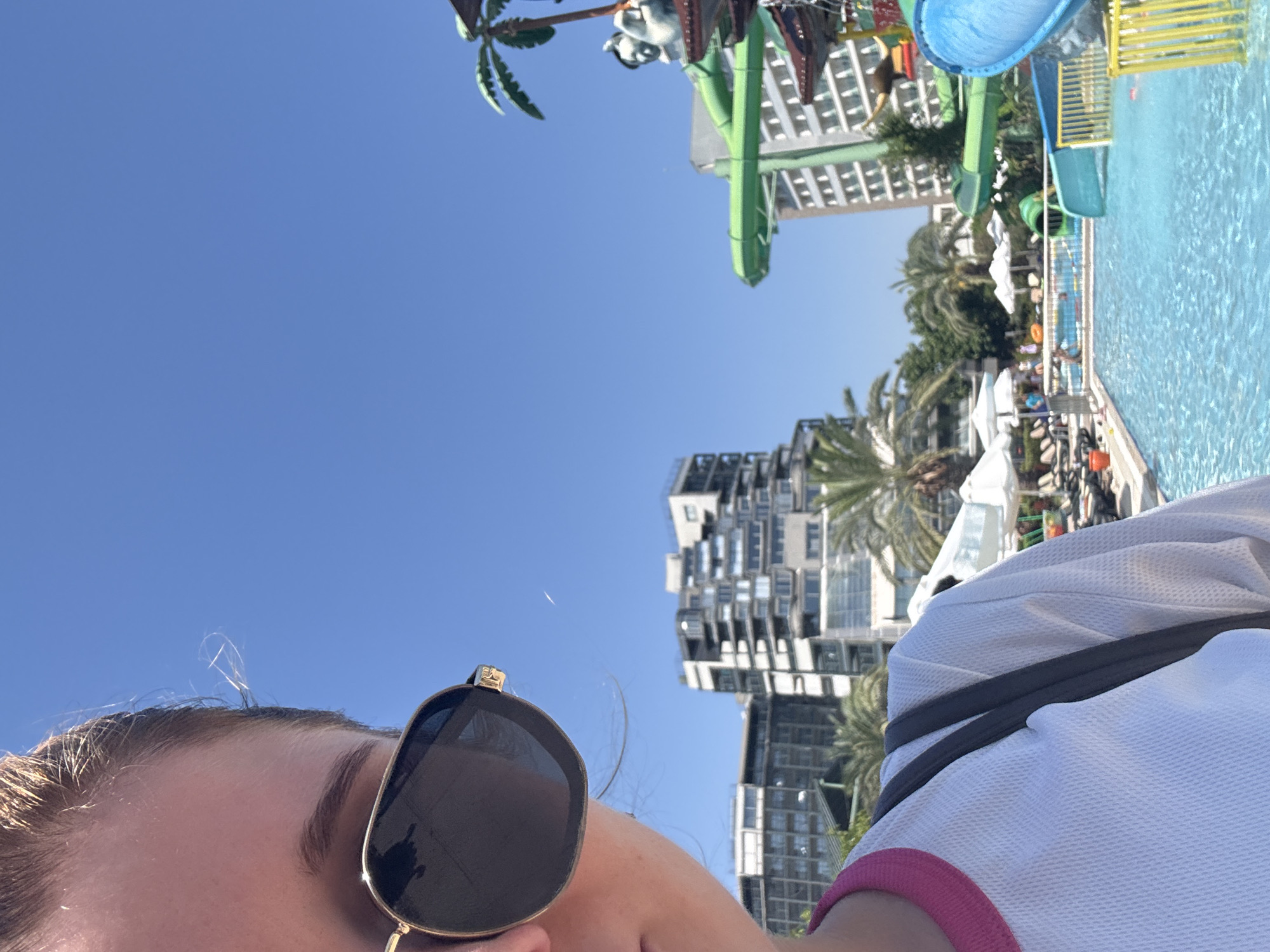 A partial selfie shows a woman wearing sunglasses and a white t-shirt with a pink collar, with a blue sky in the background. To the right, a water park is visible with green and blue slides, palm trees, and buildings. The location is Aksu, Turkey, as indicated by the presence of a water park and resort-style architecture.