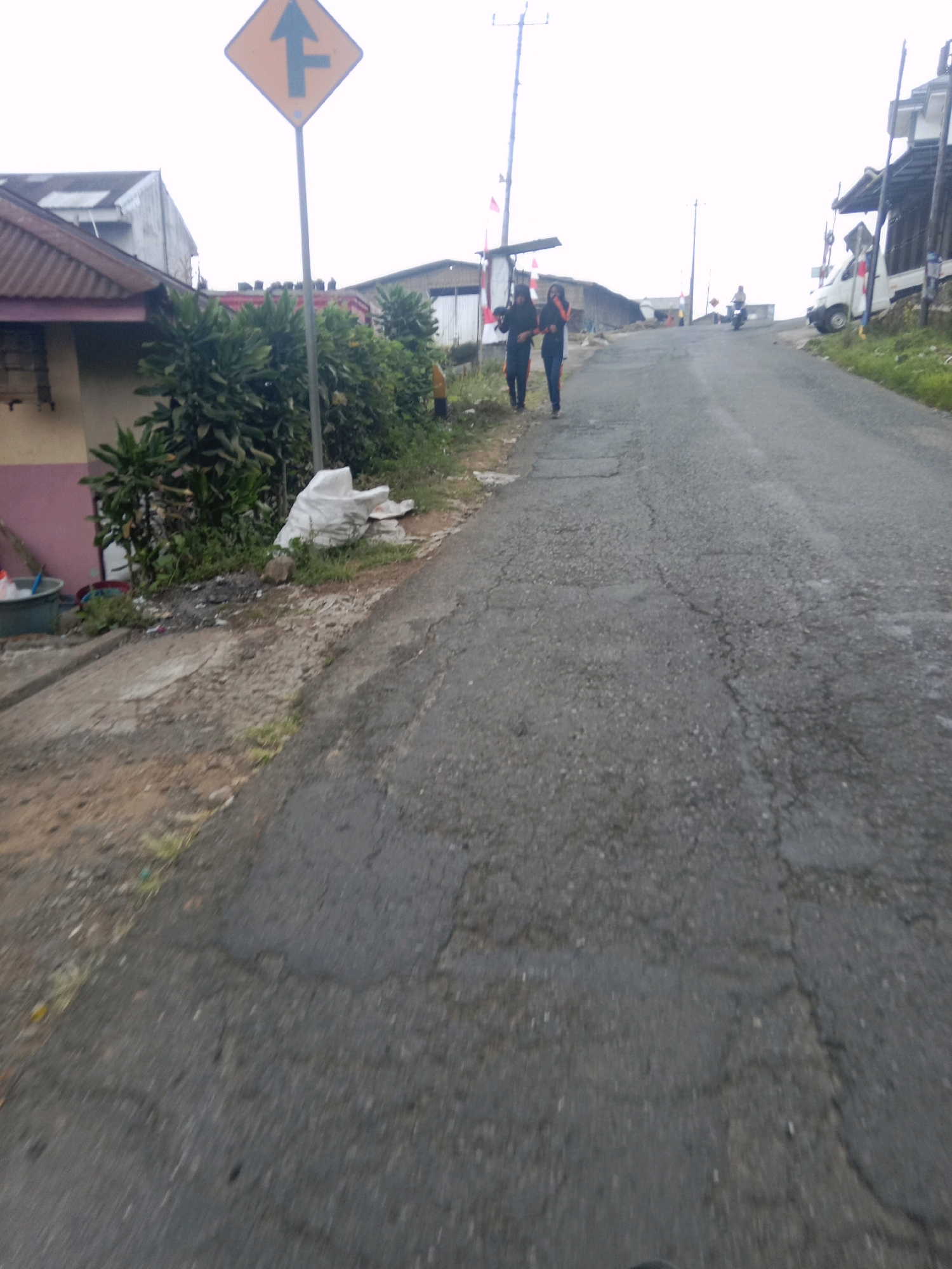 An overcast, possibly foggy, day in Batur, Indonesia, depicts a paved road with cracked asphalt, leading uphill. Two women, dressed in dark clothing with lighter colored accents, walk up the road from the left side. To the left of the road, there is a small building with a corrugated metal roof and a pinkish-tan exterior wall, surrounded by dense green foliage. A yellow diamond-shaped traffic sign with a black arrow indicating a lane merge to the right is prominently displayed on a metal pole near the building. Further up the road, another person rides a motorcycle, and a white vehicle is parked on the right side. The overall environment suggests a rural or semi-rural setting within a hilly terrain.