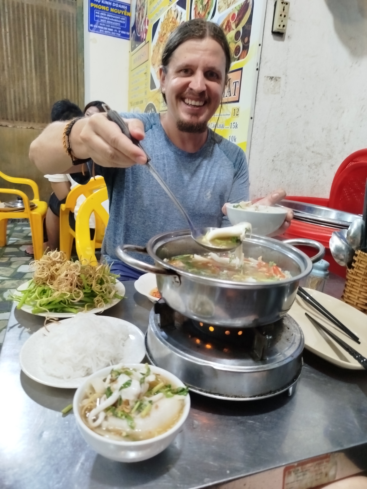 A man is eating a meal at a restaurant in Ho Chi Minh City, Vietnam. He is smiling and holding a ladle with food over a metal pot set atop a burner. The table is cluttered with food, including a plate of vegetables and noodles, as well as bowls filled with a soup or broth. In the background, other people are seated at nearby tables. There are Vietnamese characters visible on a sign on the wall. The setting appears to be a casual, local eatery.
