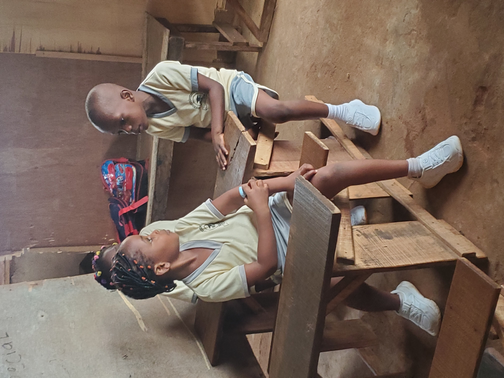 Two children wearing matching light-colored shirts, grey shorts, and white sneakers are interacting with wooden desks. One child stands on top of a desk while the other child sits on one. A red and blue backpack sits nearby. The photograph was taken indoors in what appears to be Ukwu-Abwa, Nigeria.