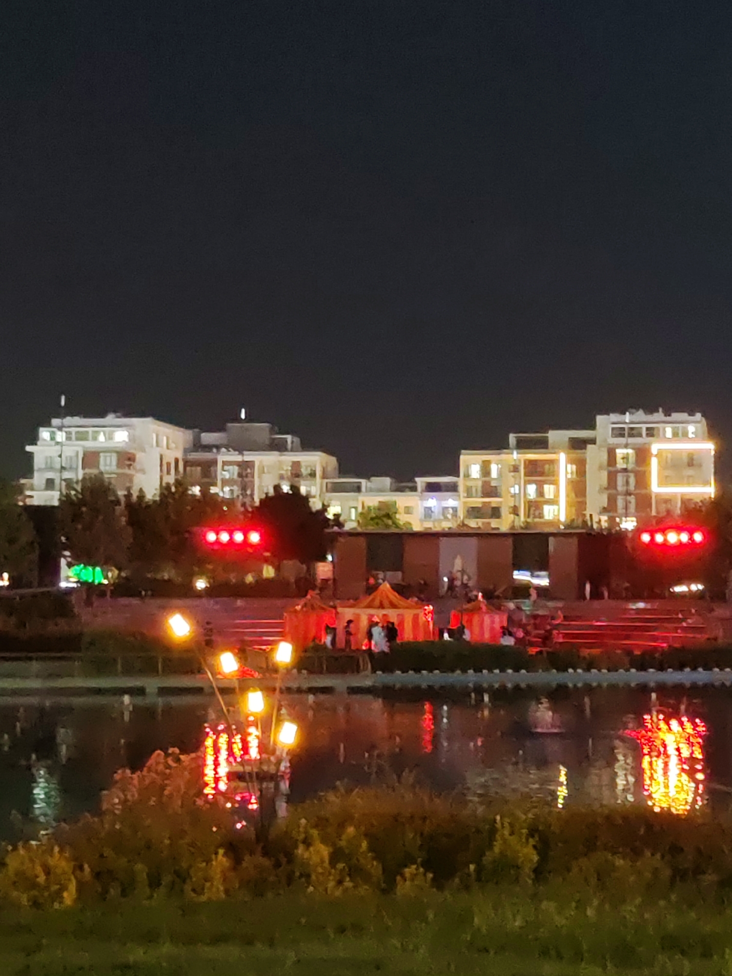 The image captures an evening scene at "Tashkent City" park in Tashkent, Uzbekistan. In the background, modern multi-story buildings with illuminated windows are visible against a dark sky. In the middle ground, a tent-like structure, possibly a stage or a decorative setup for an event, is illuminated with red and orange lights. There are several people in the vicinity of this structure. In the foreground, a body of water reflects the lights from the park. Several outdoor lamps with multiple glowing yellow bulbs are also present in the foreground. The overall impression is of a well-lit urban recreational space at night.