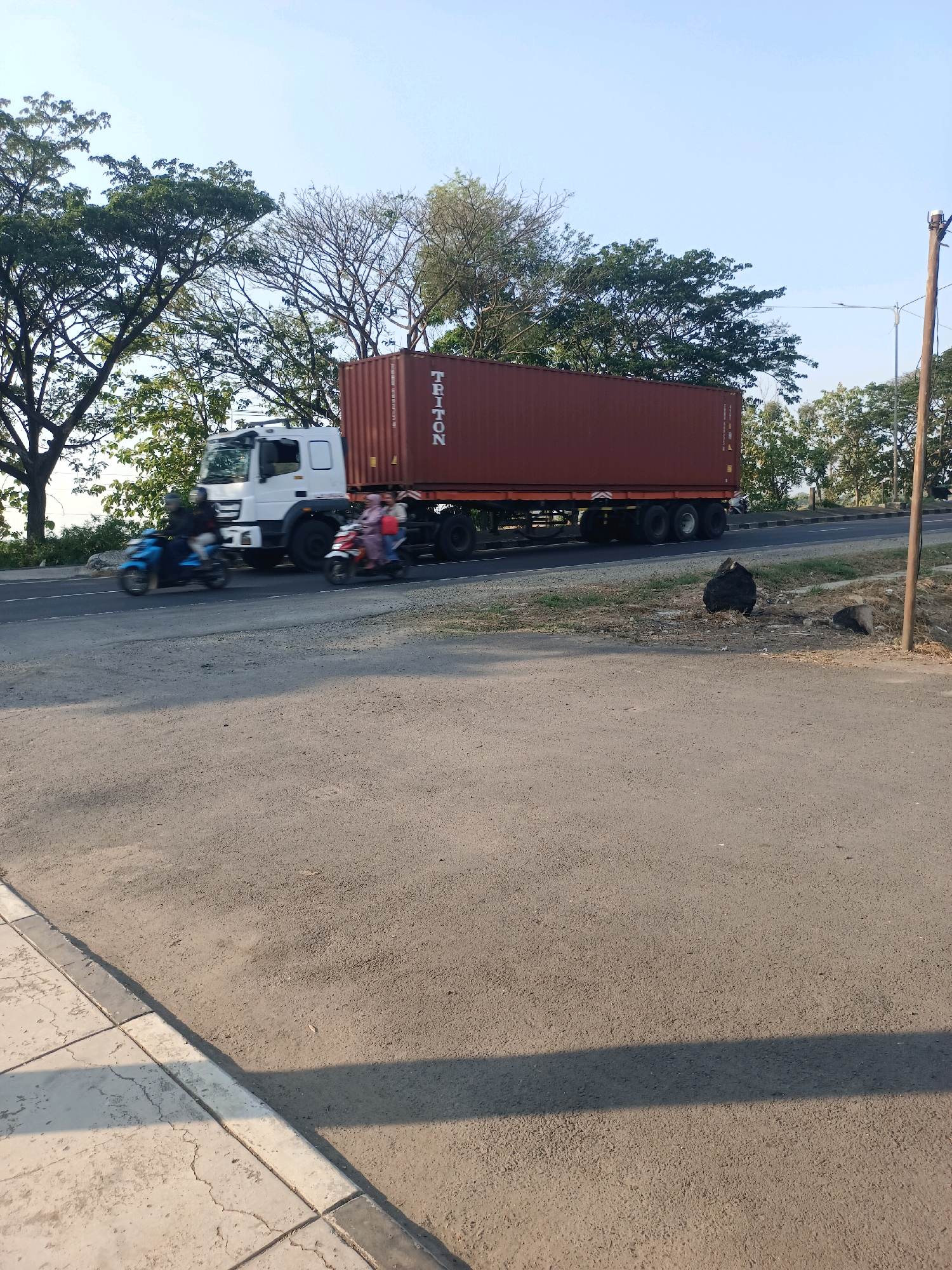 In Subang, Indonesia, a white semi-trailer truck carrying a large red shipping container moves along a paved road. The container has "TRITON" printed vertically on its side. Two motorcycles are also traveling on the road. The first motorcycle, blue, carries a single rider wearing a helmet. The second motorcycle, red, has two riders, both wearing helmets, with one appearing to be female in a pink headscarf and light-colored clothing. Trees with green foliage line the left side of the road, and a wooden utility pole stands on the right. The sky is clear and blue. The foreground consists of a paved road and a sidewalk with concrete slabs. The scene appears to be on the outskirts of the city, with a rural or semi-rural setting indicated by the vegetation and open space.