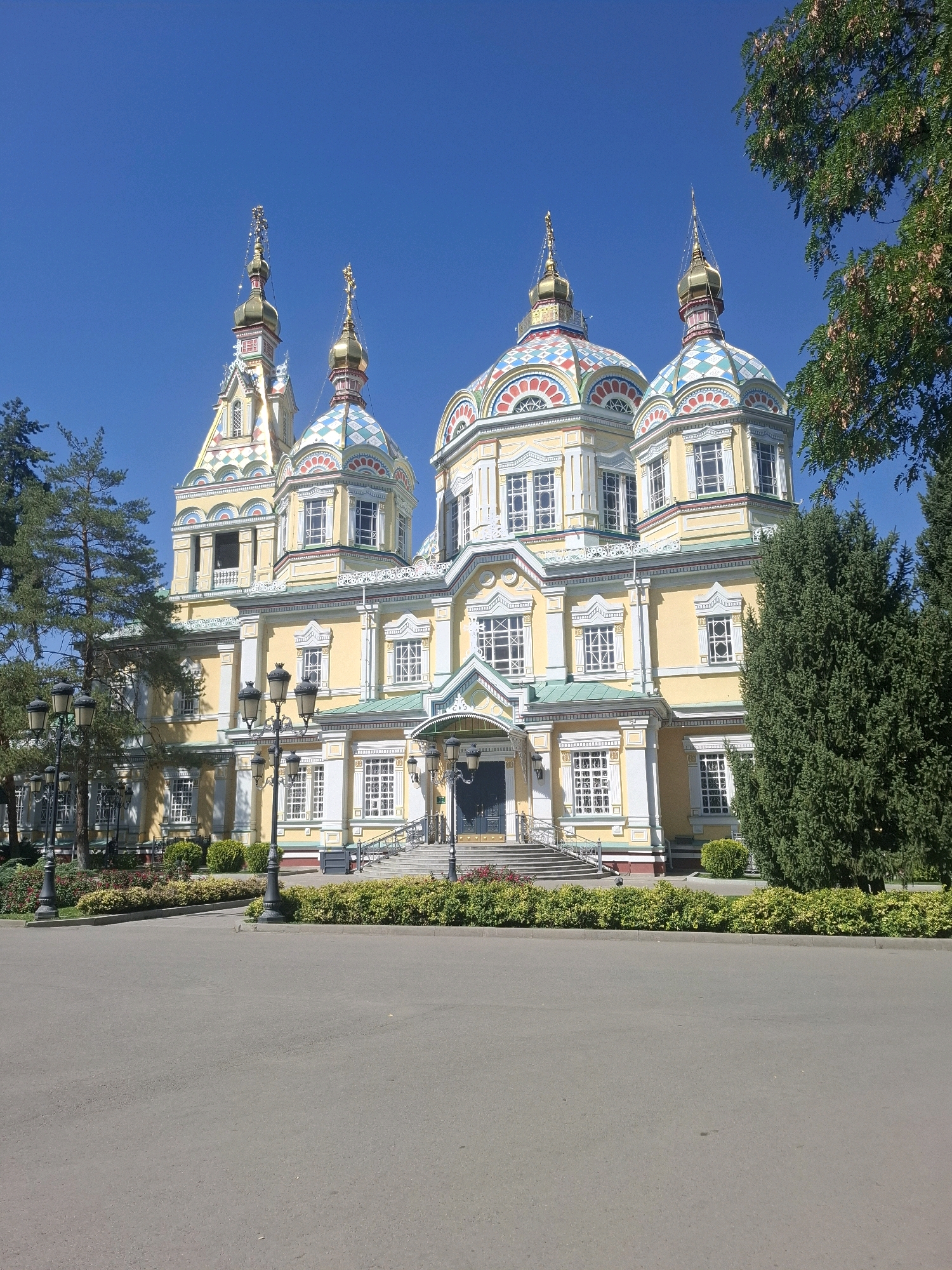 The image depicts the Zenkov Cathedral, also known as the Ascension Cathedral, located in Panfilov Park in Almaty, Kazakhstan. The cathedral is a striking example of Russian Orthodox architecture, constructed entirely of wood without the use of nails. It is painted a vibrant yellow with white trim and features multiple domes topped with golden crosses. The domes are adorned with colorful patterns and intricate detailing.
In the foreground, a paved walkway or road stretches across the bottom of the frame. To the left and right of the walkway, manicured green hedges and flowerbeds are visible. Several ornate black lampposts with multiple bulbs are positioned along the walkway, lining the entrance to the cathedral.
The cathedral itself is the central subject, a multi-story structure with numerous windows. A prominent entrance with stairs leads up to the main doors. The sky above is a clear, bright blue, indicating a sunny day. Lush green trees, including pine trees and a large evergreen, frame the sides of the cathedral and the park. The overall scene is serene and picturesque, highlighting the beauty of the cathedral and its surrounding park. No people are visible in the image.