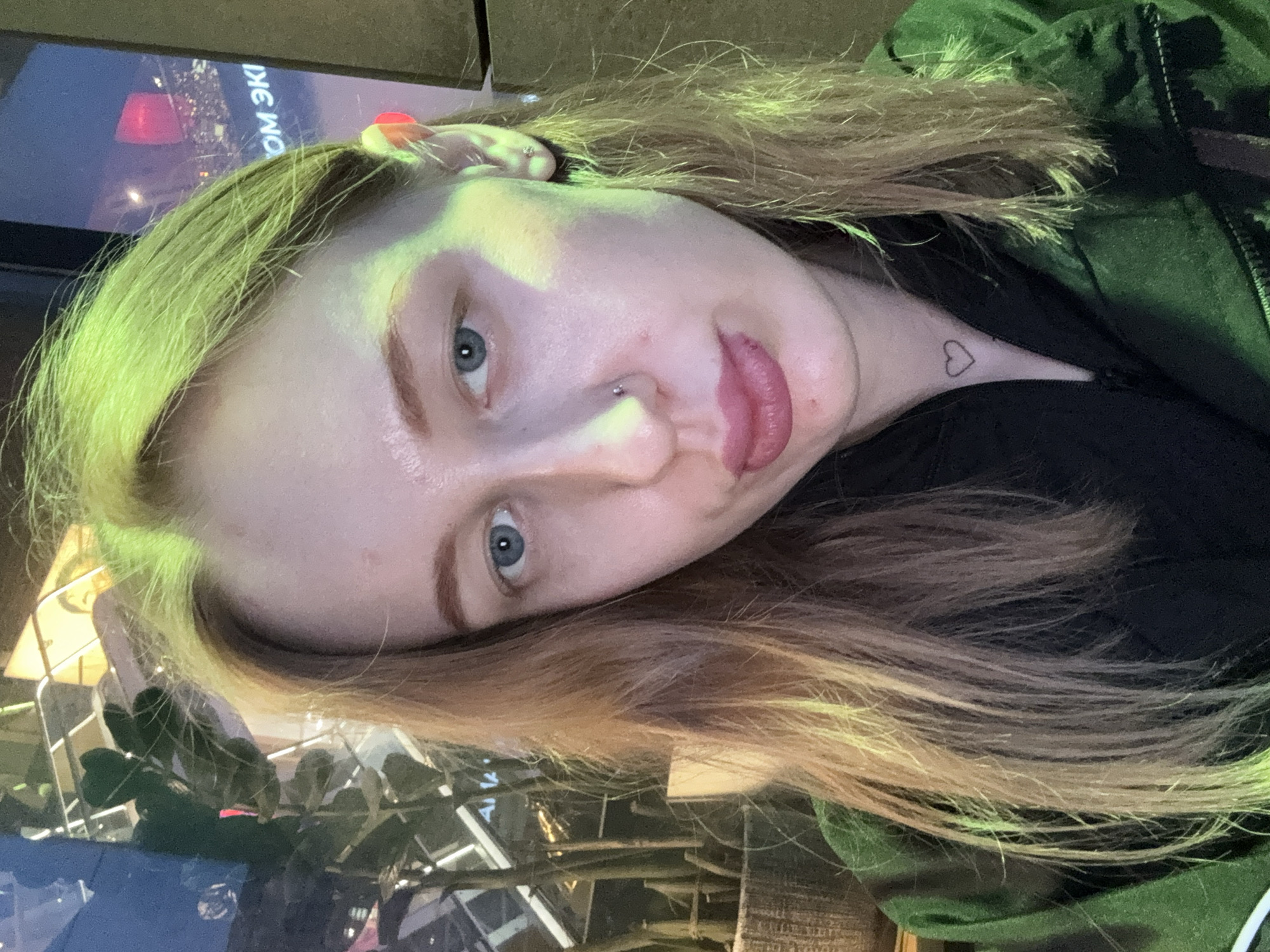 A woman is captured in a close-up shot, slightly tilted. Her hair is blonde with hints of green. The woman has fair skin and blue eyes, with a small stud piercing on her nose. A small heart tattoo is visible on her neck. She is wearing a black shirt or top underneath a green jacket.
The background includes a blurred screen or display with text in a Cyrillic alphabet. There is also vegetation visible, likely plants, and architectural elements that suggest an indoor space, possibly a restaurant or public venue. The image was taken in Ol'ginskaya, Russia.
