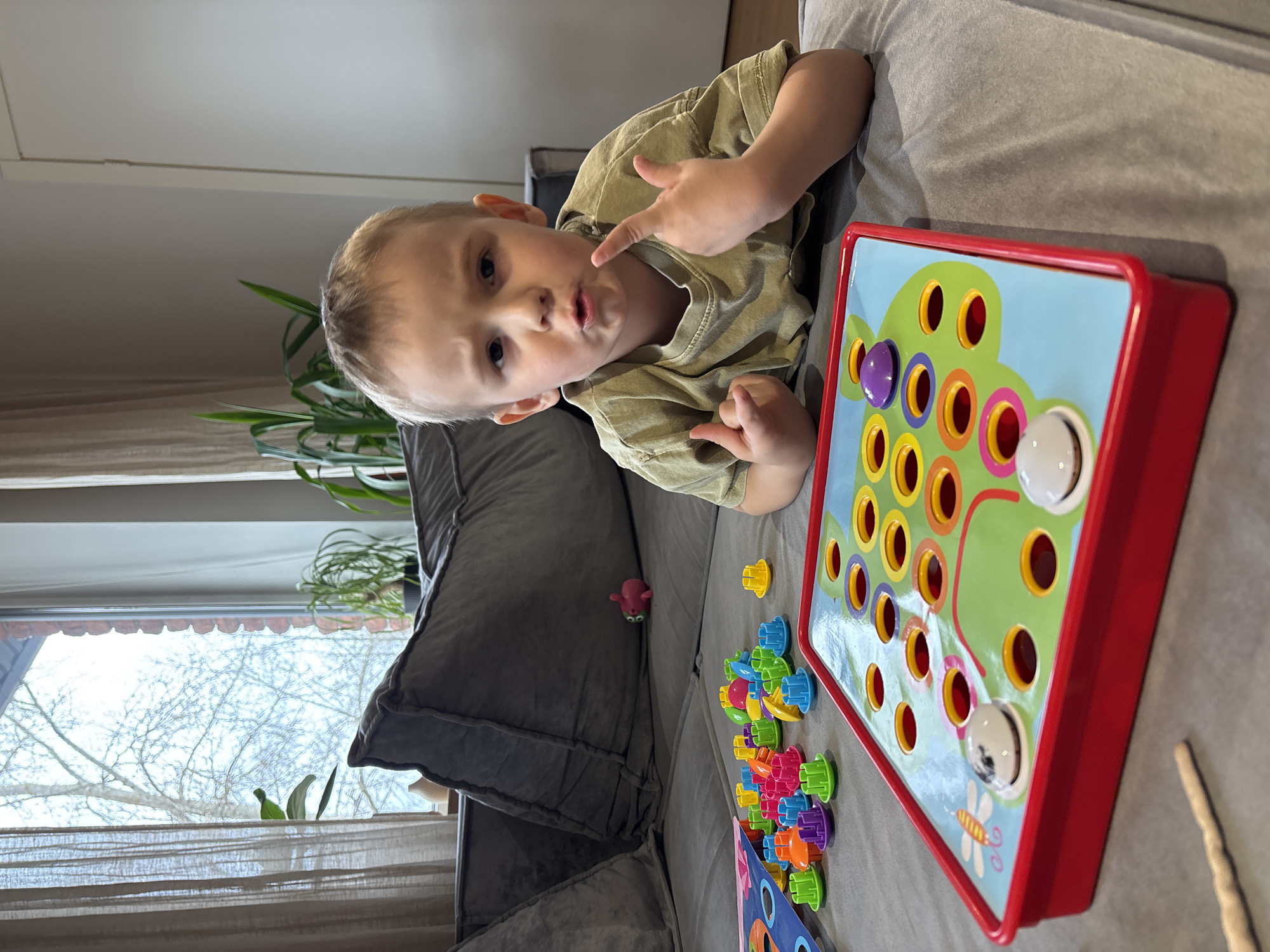 A young boy is lying on a gray couch, playing with a colorful toy featuring holes and pegs. The boy is wearing a light green shirt and pointing with his right index finger towards his mouth. The toy, positioned on the couch next to him, has a red frame, a light blue background, and a design of colorful circles and a green path. Various colorful pegs are scattered around the toy on the couch. A window with bare trees visible through the partially drawn curtains is in the background. The scene appears to be indoors, possibly in a living room. There is no evidence to suggest that this photo was taken in Chaikovsky, Russia.