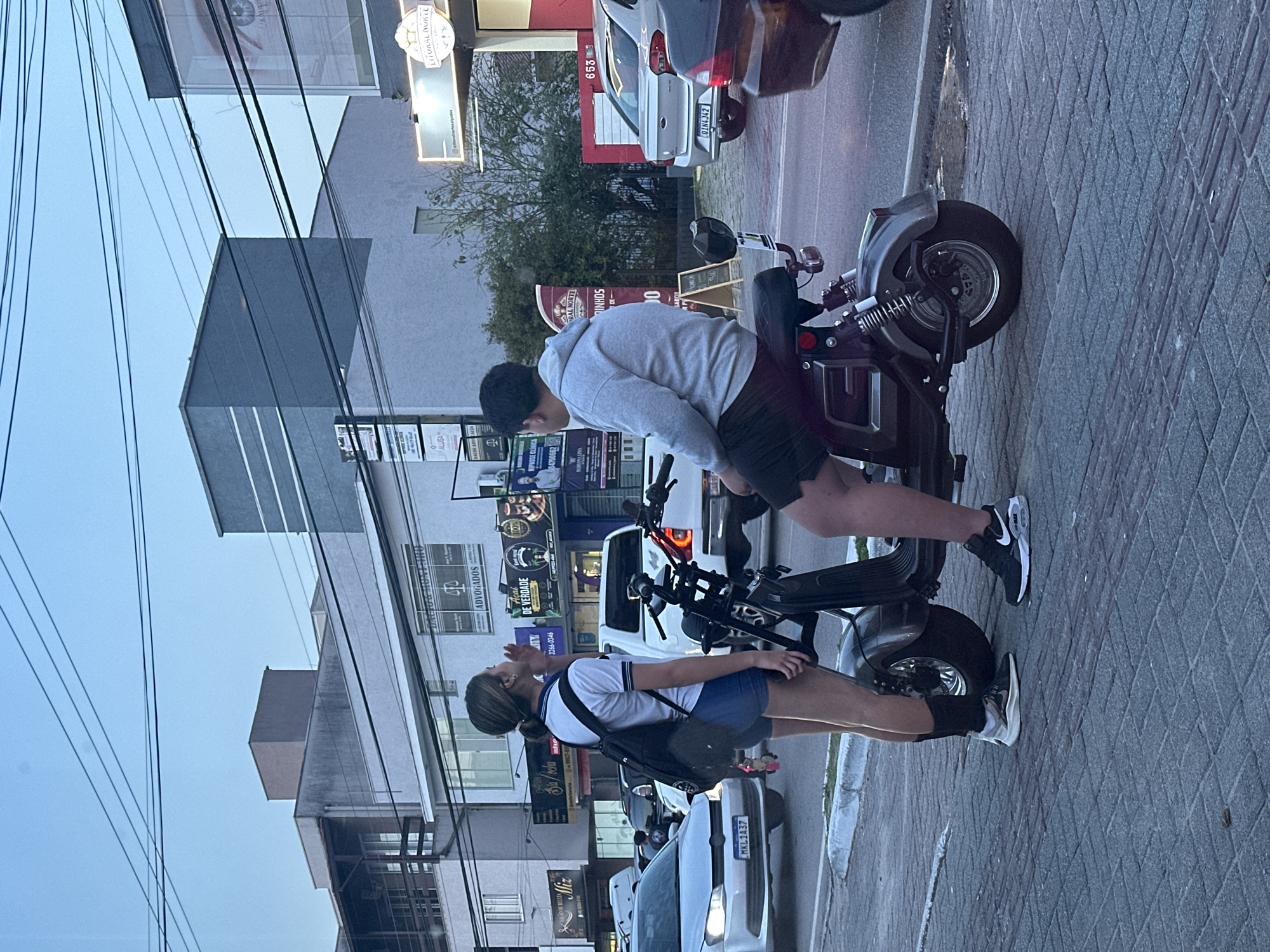 In Florianópolis, Brazil, a young woman stands on a paved sidewalk next to an electric scooter. She is wearing a white polo shirt, blue shorts, black socks, and athletic shoes. A black backpack is slung over her shoulders. She appears to be speaking or gesturing with her right hand near her mouth. A young man sits on the electric scooter, facing away from the camera, wearing a grey hooded sweatshirt and dark shorts. His legs are visible from the knees down, and he is wearing black athletic shoes. The scooter is dark grey with chrome accents and a red taillight. Behind the subjects, there is a street with several cars, including a white Volkswagen Golf with license plate MKL1A37 and a silver Ford Ka with license plate OIN4J42. Commercial buildings line the street, featuring signage for various businesses, including "ADVOGADOS," "HIPNOSE CLÍNICA," and "LITORAL NORTE." The sky is overcast and appears to be dusk. Overhead, power lines crisscross the frame.