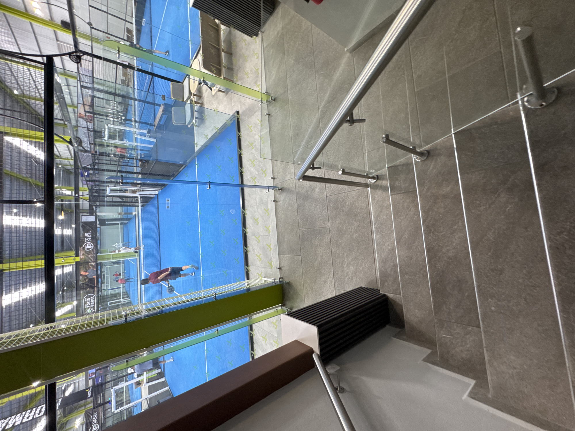 This image captures an indoor padel tennis facility, viewed from an elevated perspective, likely from a staircase or viewing platform. The scene is dominated by bright blue padel courts on the left and a modern staircase with railings on the right.
On the left side of the frame, several padel courts are visible. Each court features a vibrant blue playing surface with clear white lines, enclosed by transparent glass walls and black metal mesh fencing. Multiple individuals are engaged in play or observation on these courts. One player, prominently featured in the mid-ground, wears a dark red shirt and shorts, holding a padel racket, and appears to be in motion. Other players are further in the background on adjacent courts.
To the right, a contemporary staircase descends, featuring dark grey textured tiles on the treads and risers. A sleek handrail system runs alongside the stairs, comprising polished silver metal bars and clear glass panels, providing both support and an unobstructed view of the courts. A dark, ridged structure, possibly a bench or architectural element, is situated next to the stairs.
The facility's ceiling is high, with an industrial design featuring metal beams and corrugated panels, allowing natural light to filter in through skylights. Various banners and signs are displayed around the courts. Visible text includes "TOUR PADEL" and "TSAL" on banners, and "OHMA" (likely part of "OHMAPADEL") on a sign near the bottom left. Green, Y-shaped patterns are also visible along the light green border of the court structures. The overall impression is a busy, well-lit, and modern sports complex during the daytime in San Bernardino Tlaxcalancingo, Mexico.