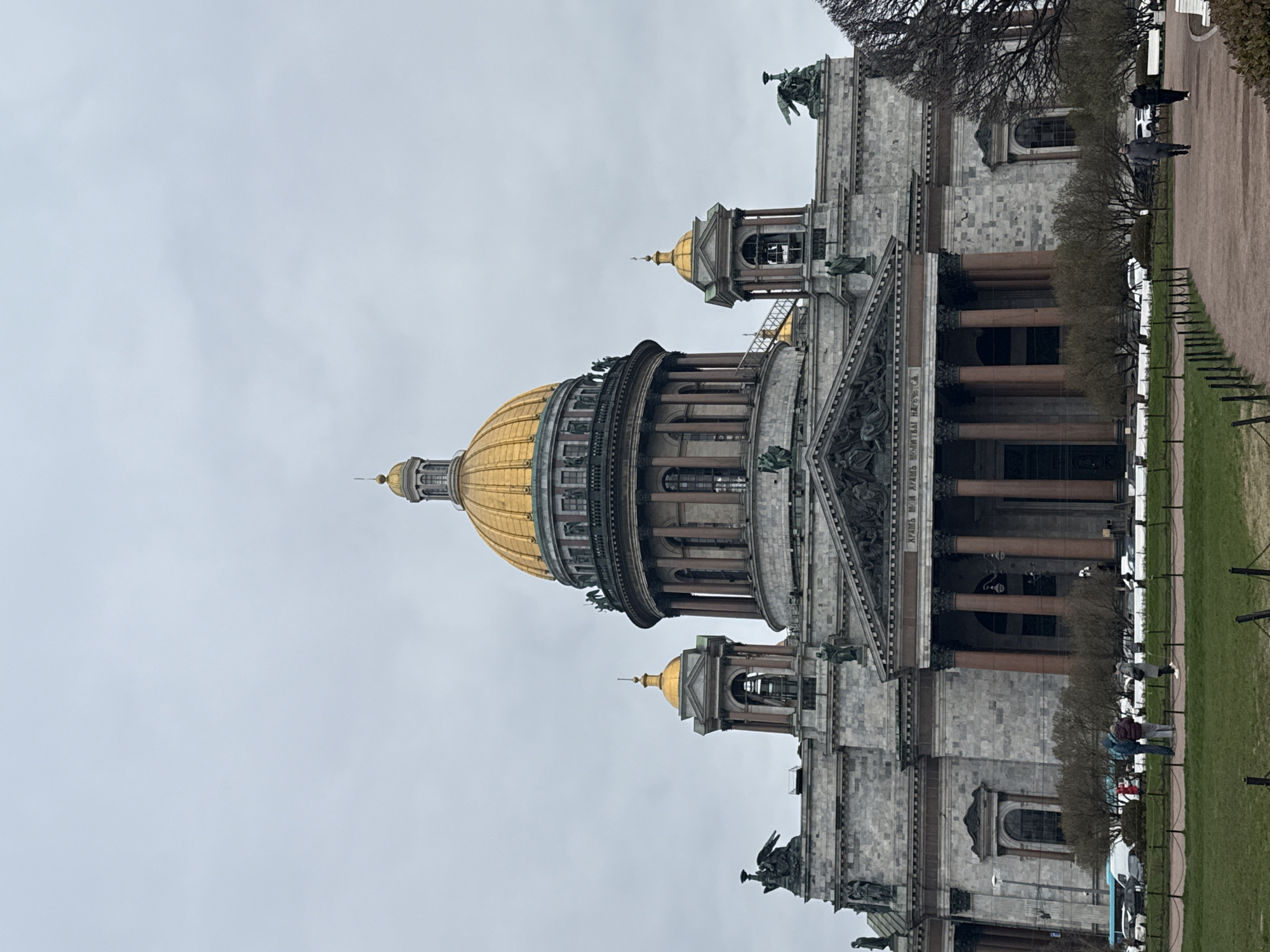 This media file presents a grand, neoclassical cathedral, identified as St. Isaac's Cathedral in Saint Petersburg, Russia, under an overcast sky. The magnificent building occupies the left side of the frame, showcasing a prominent central golden dome and several smaller golden cupolas and bell towers. The cathedral's exterior is constructed from light-colored stone, adorned with numerous large columns and intricate sculptural details, including statues along the roofline and relief carvings above the main entrance portico.
To the right of the cathedral, there is a paved path alongside a grassy area, separated by a low fence. Several people are visible on the path, walking or standing casually. The trees adjacent to the path have bare branches, suggesting the time of year is late winter or early spring. The sky is uniformly light grey, indicating an overcast daytime.
A significant detail is the inscription visible on the frieze above the main portico, which reads in Cyrillic: "ЦАРЮ МОЕМУ И БОГУ МОЕМУ". This translates to "To my King and my God." The scene captures a tranquil moment at this iconic architectural landmark, with a few visitors observing the surroundings.