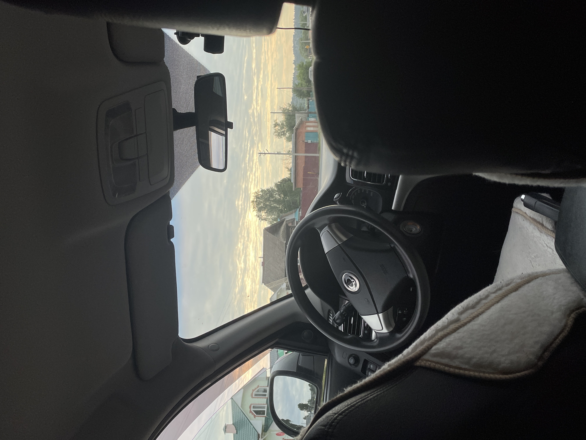 The image shows the interior of a car looking out at a rural scene in Gavrilovka, Russia, during what appears to be sunrise or sunset. The view from the driver's seat includes the steering wheel, dashboard, and side mirror reflecting the sky and trees. Outside the car, there are houses with green roofs, brick buildings, and power lines against a cloudy sky with warm hues. The sun is low on the horizon, casting a soft light on the landscape. There are no people visible in the image.