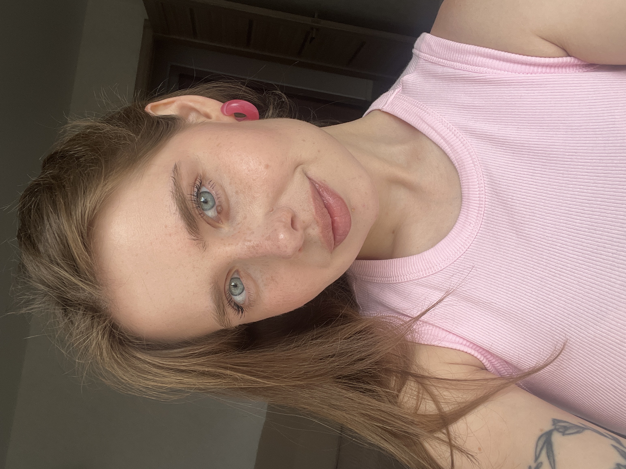 A young woman with blonde hair and blue eyes is shown in a close-up selfie. She is wearing a pink ribbed tank top and a pink hoop earring. Her hair is pulled back, with some strands falling around her face. She has freckles across her nose and cheeks. The background is blurred, but appears to be an indoor setting with a door visible on the right. The image was taken in Chaikovsky, Russia.