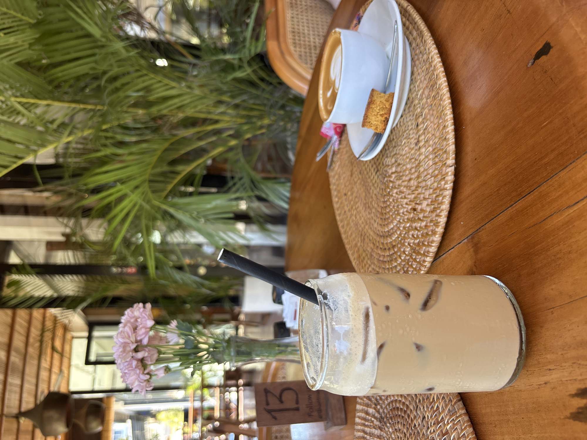The image captures a table scene in Pecatu, Indonesia. On a wooden table with a woven placemat, sits a glass of iced coffee with a black straw. Nearby, a small vase holds pink flowers. In the background, a woven chair and a white cup of coffee on a saucer are visible, set on another woven placemat. A small piece of pastry rests on the saucer. Palm fronds fill the upper left portion of the frame. The scene is outdoors, with building structures and natural light visible. A wooden table number stand is present.