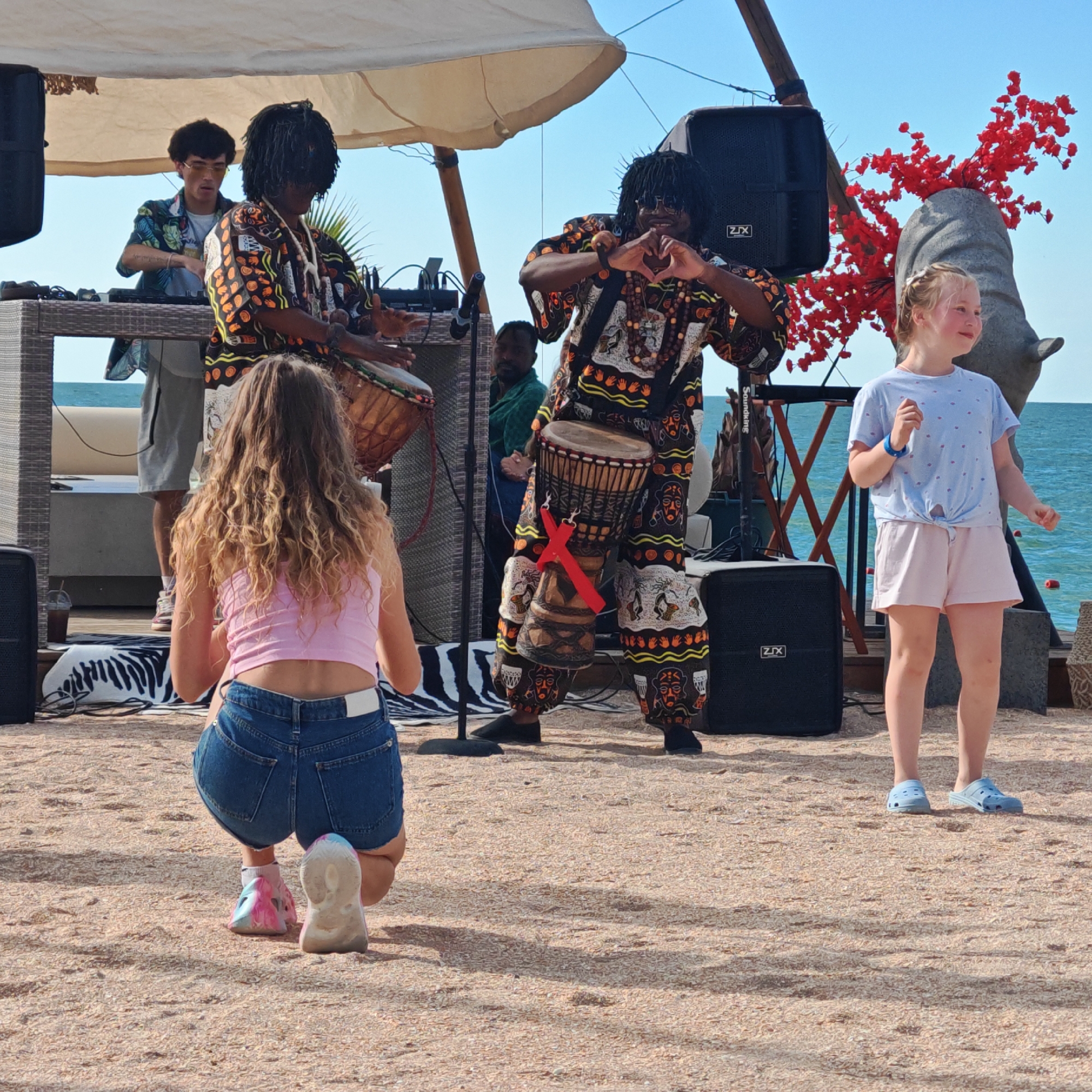 In a sandy outdoor setting, likely near a body of water, a group of people are gathered. The focal point is a performance involving at least two musicians playing drums. One musician, wearing a vibrantly patterned outfit, is actively playing a drum and making a heart shape with his hands. Another musician, in the background to the left, is also playing a drum. A younger girl in a pink top and denim shorts is crouched in the foreground, facing away from the camera, seemingly observing the performance. To the right, another young girl in a light blue t-shirt and pink shorts stands and watches. Behind the performers, a stone sculpture of an animal's head adorned with red decorative branches is visible, along with large speakers and part of a tent structure. The presence of speakers and what appears to be a DJ setup suggests a musical event or performance. The background reveals the sea and a clear sky.