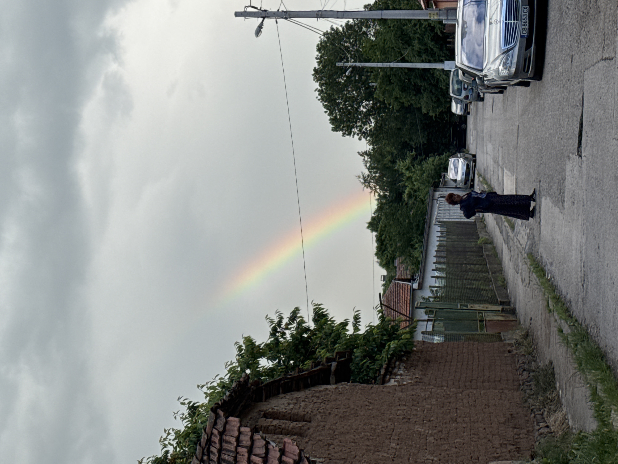 The image depicts a residential street scene in Sofia, Bulgaria, under an overcast sky with a rainbow visible.
The upper portion of the frame is dominated by a heavy, light grey cloud cover. A distinct, partial rainbow arcs across the sky from the left to the upper center, displaying the spectrum of colors.
On the left, a textured, reddish-brown mud brick or stucco wall belonging to an old building occupies the foreground, topped with weathered red-orange roof tiles. Lush green foliage, likely from a tree or large bush, spills over the wall and roofline. Beyond this wall, another house with a tiled roof is partially visible.
The street, a paved asphalt surface, runs from the foreground into the background, showing cracks, patches, and potholes. A concrete sidewalk, partially overgrown with grass, parallels the wall on the left.
On the right side of the street, a woman with reddish-brown hair is standing, facing slightly to her right (viewer's left). She wears a dark, long-sleeved top and a floor-length dark skirt or dress, with sneakers on her feet. Her left hand is placed on her hip.
Several cars are parked along the right side of the street. In the immediate foreground, a silver Mercedes-Benz S-Class sedan (W221 generation) is parked facing the viewer, its front grille and headlights prominent. The vehicle bears a Bulgarian license plate "CB 8653 CX," with "CB" identifying it as registered in Sofia. Behind the Mercedes, other vehicles, including a light-colored hatchback and a grey SUV or wagon, are visible further down the street.
A grey utility pole with a street lamp and multiple power lines extends vertically on the right side of the frame. Densely packed green trees line both sides of the street further into the distance, with additional houses partially visible beyond the tree line. The overall environment suggests a somewhat older, possibly suburban or semi-rural residential area within Sofia.