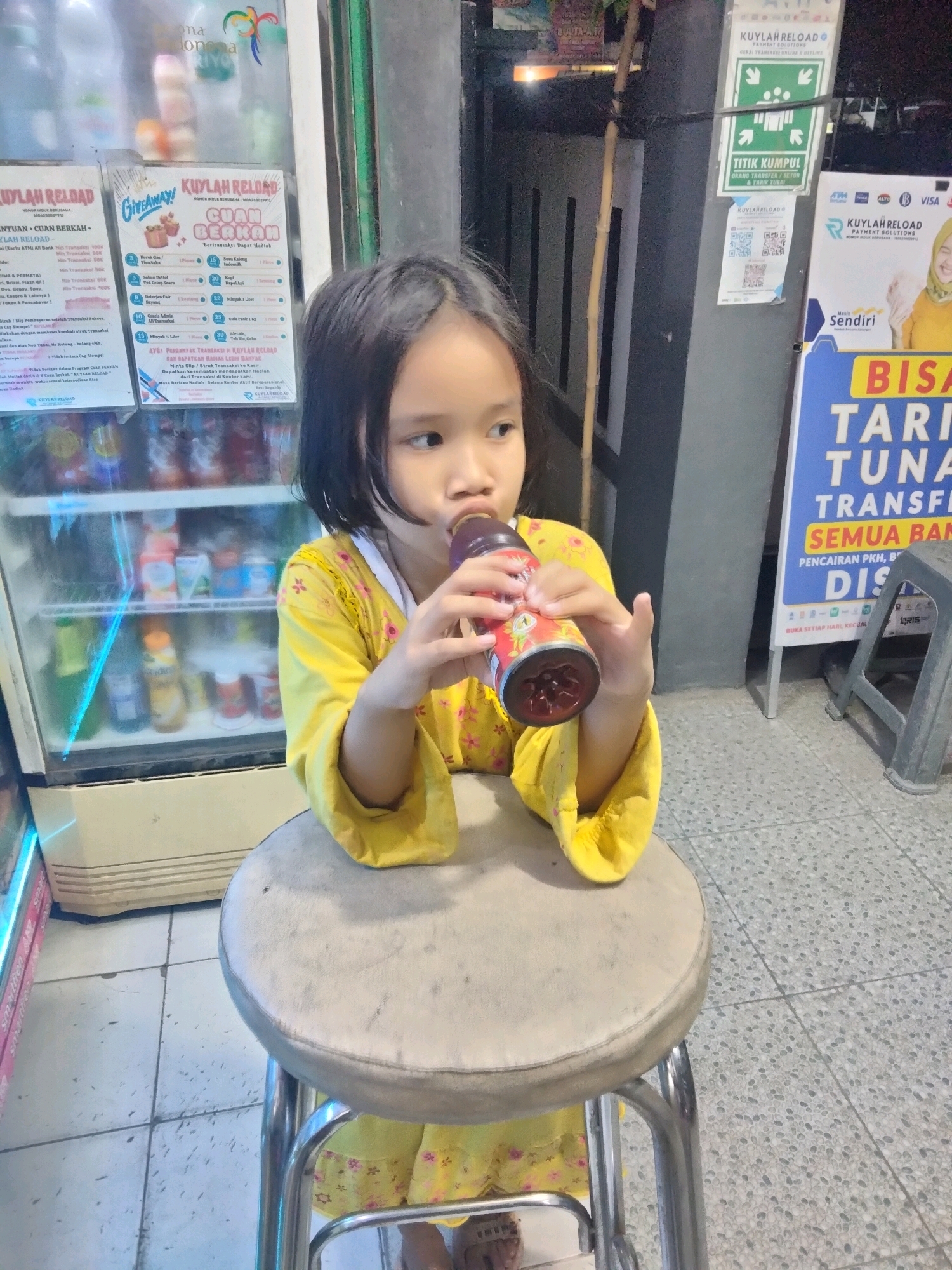 A young Indonesian girl with dark hair and fair skin is sitting on a stool in front of a refrigerator and a commercial sign. She is wearing a bright yellow dress with a floral pattern. The girl is drinking from a dark-colored bottle, holding it with both hands. The refrigerator behind her is filled with various drinks. The commercial sign to her right features text in Indonesian, including "KUYLAR RELOAD" and "BISA TARIK TUNAI". The floor is tiled with a speckled pattern. The overall scene appears to be an outdoor or semi-outdoor setting, possibly a small shop or kiosk in Suka Sari, Indonesia.