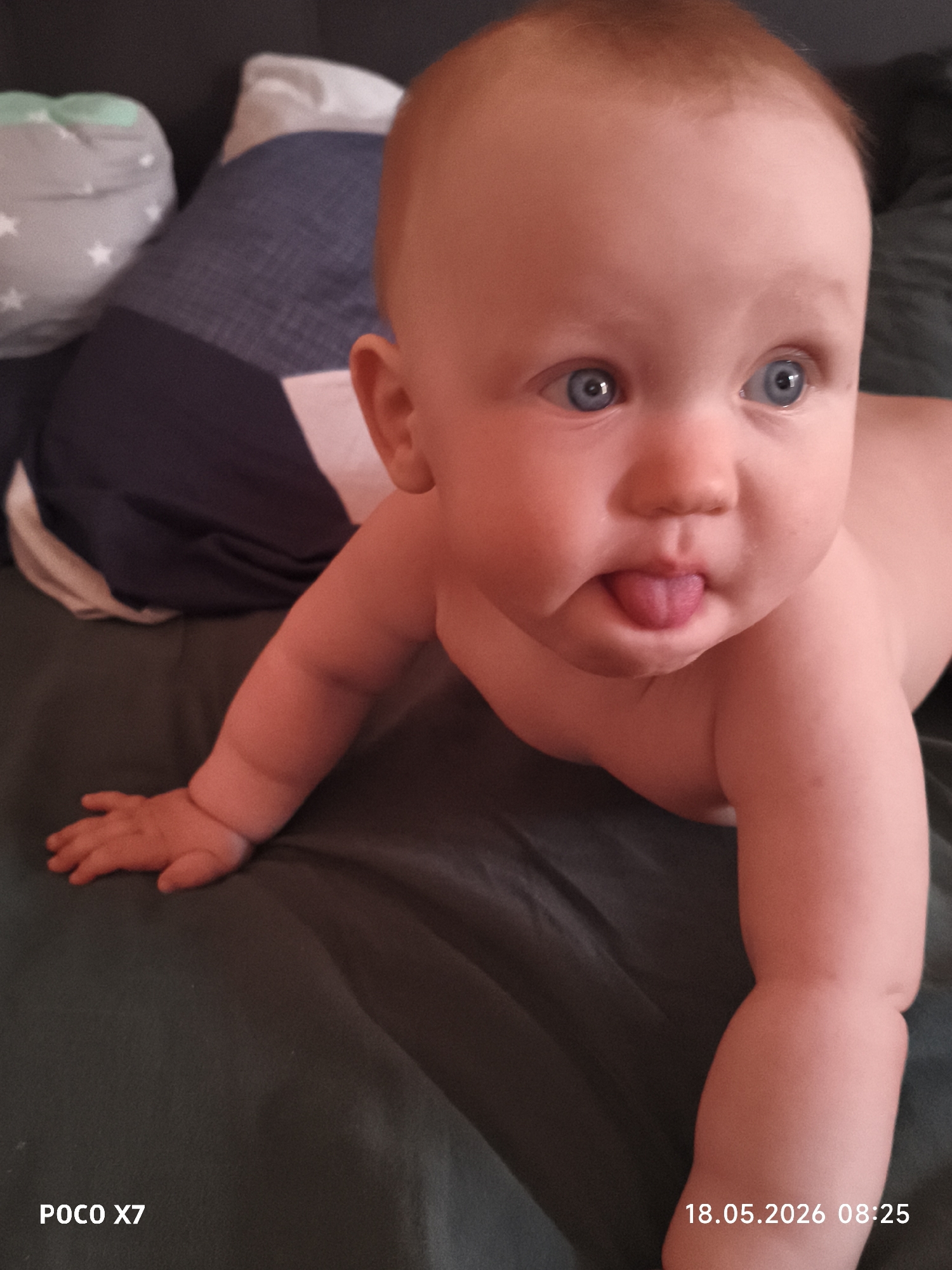 A baby with blue eyes and red hair is lying on their stomach, sticking out their tongue. The baby is on a dark green sheet with a pillow behind them. The image was taken with a POCO X7 camera on May 18, 2026, at 08:25. The location is not provided in the image.