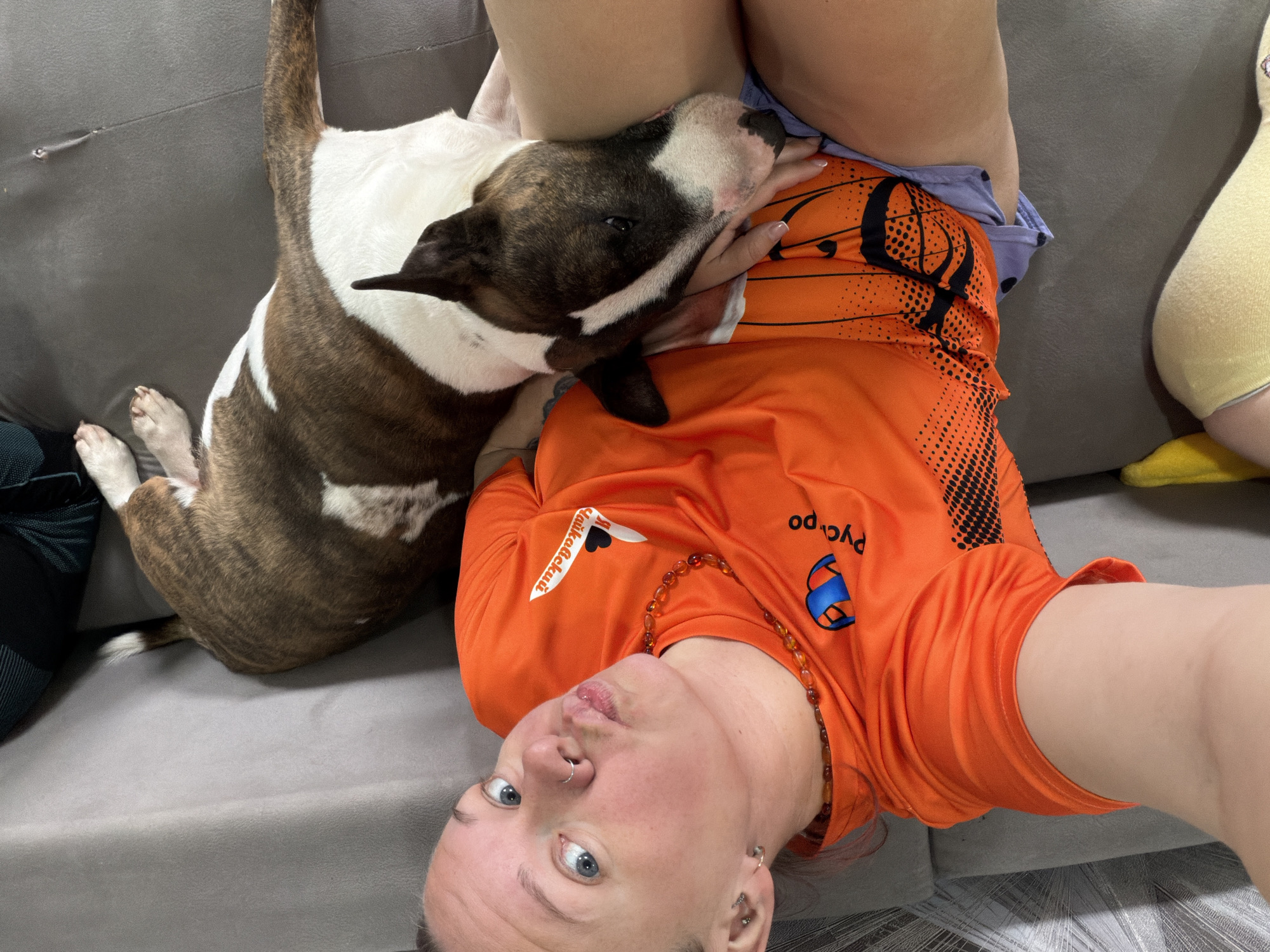 The image depicts a woman and her brindle and white Bull Terrier dog sharing a calm, affectionate moment indoors on a gray fabric sofa. The woman is lying on her back, looking directly into the camera, suggesting she is taking a selfie. She has blue eyes, a nose ring, and several ear piercings, and wears an amber-colored beaded necklace. She is dressed in an orange short-sleeved t-shirt featuring a black pattern resembling a basketball, and potentially orange shorts underneath.
Her dog is comfortably nestled on her lap and torso, with its head resting against her side, looking up affectionately at the woman. The scene conveys a strong bond between the owner and her pet.
Visible text on the woman's orange t-shirt includes "Сайкавский" (Saikavsky) with a small heart icon near her left shoulder, and partially visible "Pycpo" (Ruspo) near her right hip, both in black and white lettering. The background is primarily the gray sofa. The location context provided is Chaikovsky, Russia, which aligns with the "Сайкавский" text on the shirt.