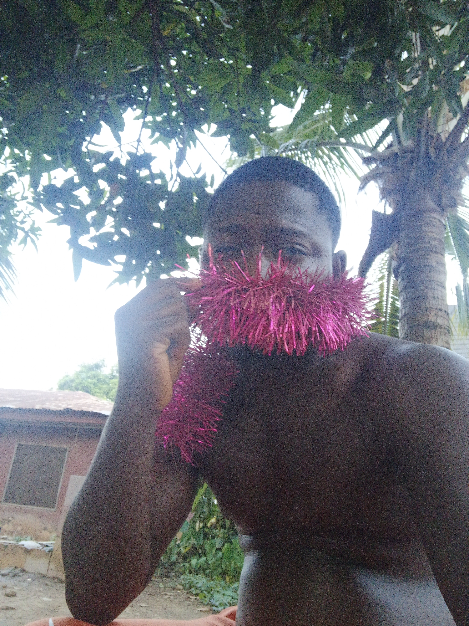 A medium shot depicts a dark-skinned, bare-chested man holding a pink metallic tinsel garland in front of his face, obscuring his mouth and nose. He is holding the garland on the left side of the frame with his left hand. The background includes a green foliage canopy overhead, a palm tree to the right, and a small building with a visible window on the left. A patch of green foliage and some ground are visible in the lower part of the frame. The location is within Akwanga, Nigeria.