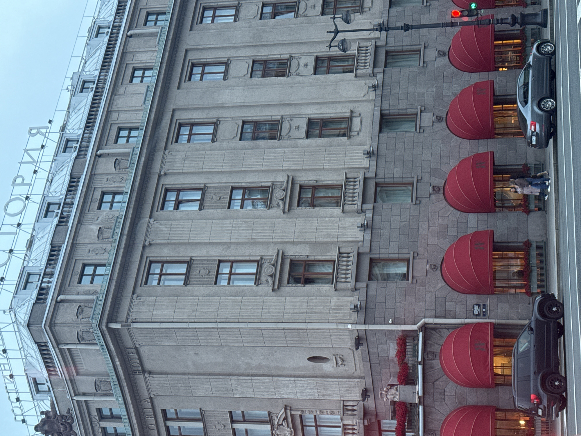The image depicts the Astoria Hotel in Saint Petersburg, Russia, on a cloudy day. The grand, historic hotel building dominates the frame, characterized by its ornate neoclassical architecture. The facade is constructed with grey stone and features multiple stories with numerous windows. Prominent red awnings are visible over the ground-floor windows, adding a splash of color to the otherwise muted tones of the building. A black SUV is parked on the street in front of the hotel, and a grey sedan is driving away. Two individuals are walking on the sidewalk near the hotel entrance. A streetlamp with multiple globes stands on the right side of the image, and a traffic light shows a red signal. The sky is overcast, suggesting a cool or damp day.