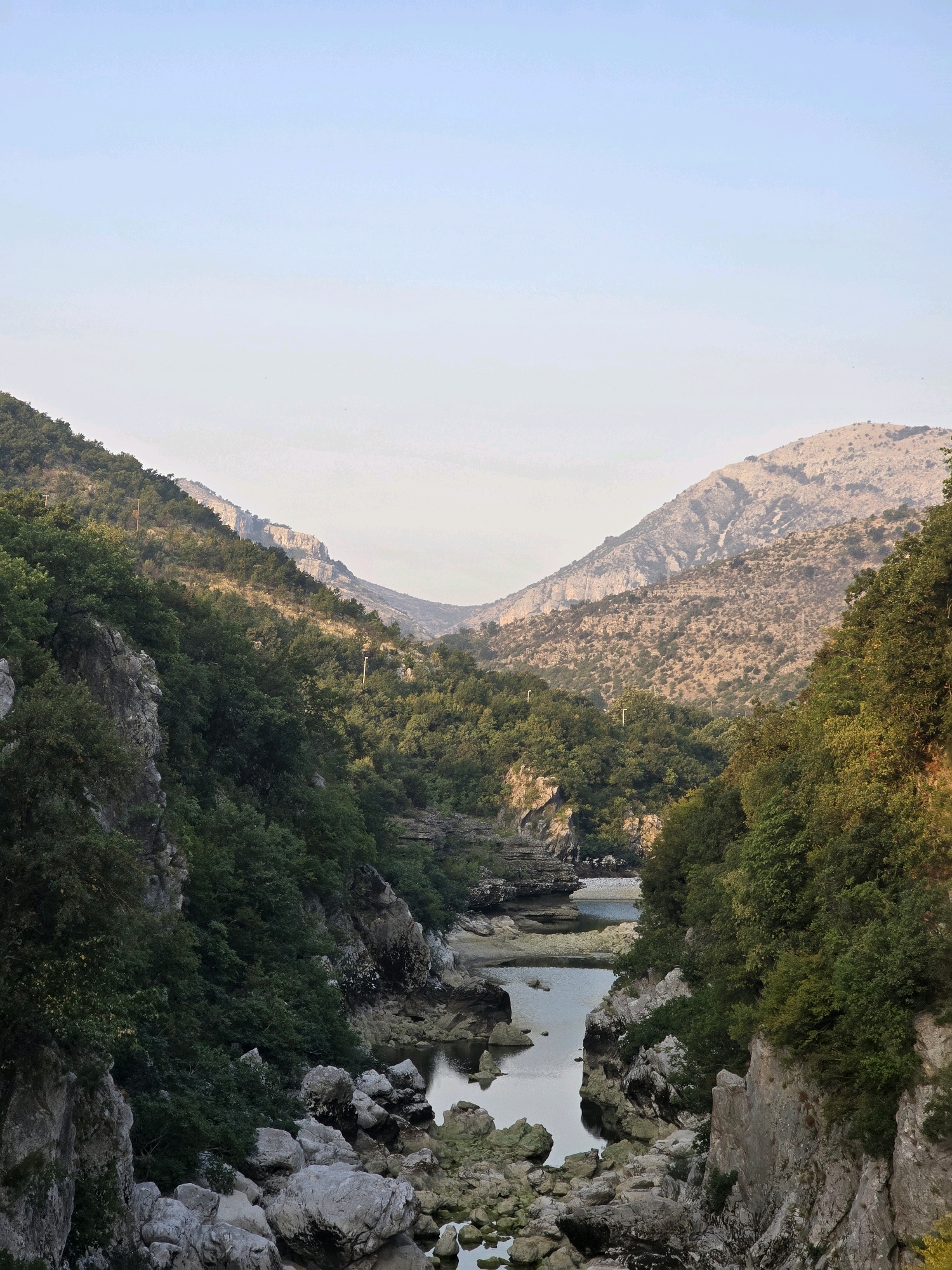 The image shows a serene natural landscape with a river flowing through a valley surrounded by lush green hills and mountains. The sky is a clear, pale blue. The foreground is dominated by the rocky banks of the river, with large grey stones scattered along the edges. The river itself is calm and reflects the surrounding greenery. Further back, the valley walls rise steeply, covered in dense foliage. In the distance, the mountains are more barren, with rocky slopes and sparse vegetation. The overall impression is one of natural beauty and tranquility. This scene is likely located in the vicinity of Bioče, Montenegro, a region known for its dramatic river valleys and mountainous terrain.