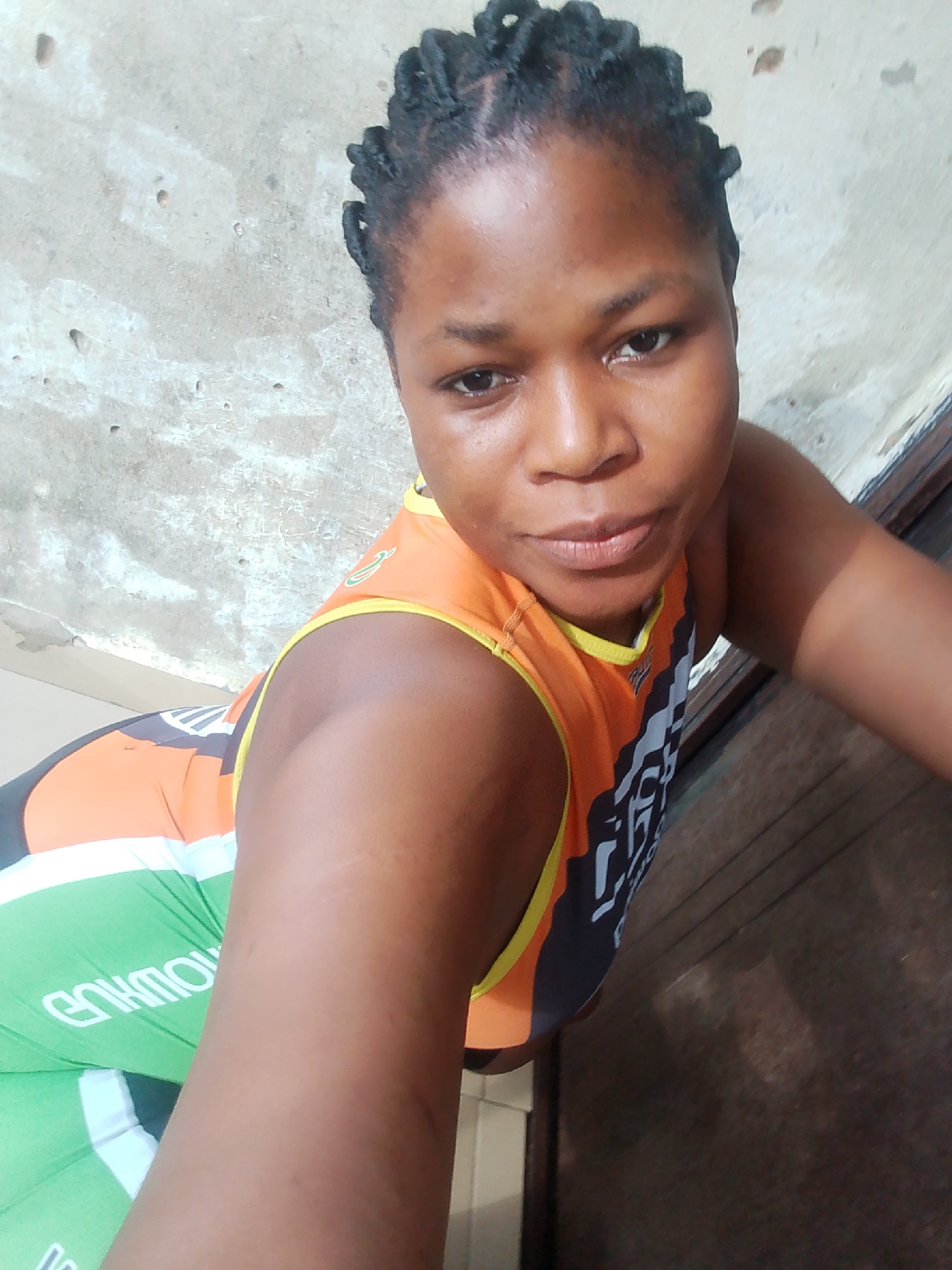 The image shows a young woman with braided hair, wearing an orange and black athletic top and green leggings. She is positioned at an angle, leaning forward and looking towards the camera. The background appears to be an indoor environment with textured walls and a wooden surface. The setting suggests a casual or athletic context, possibly in Yenegoa, Nigeria, given the prompt's request. The woman's expression is neutral, and she is the sole subject in the frame.