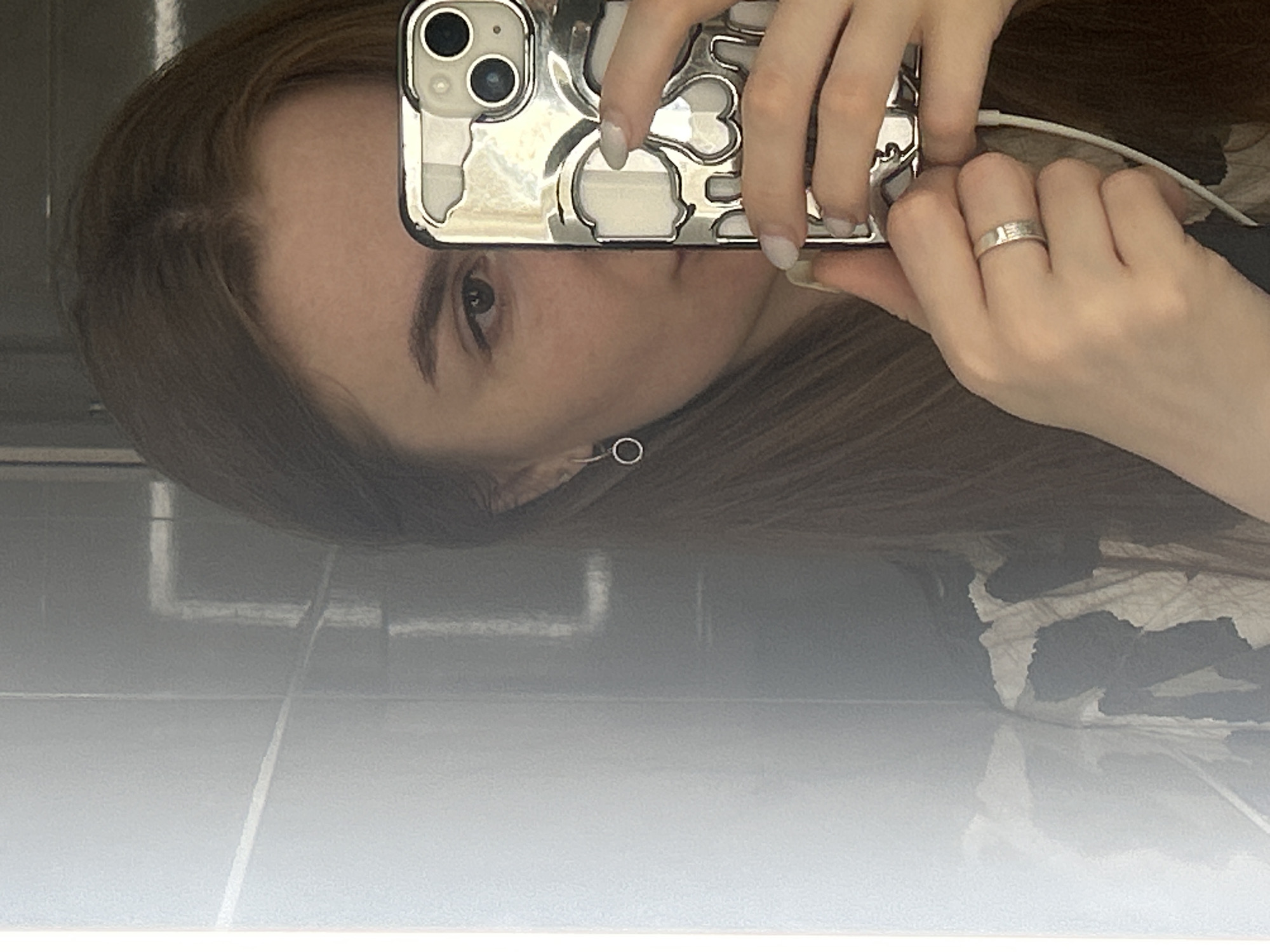 A young woman with brown hair and light skin takes a selfie in a bathroom with tiled walls. She is holding a silver phone with a skull-patterned case up to her face, covering the right side of her face. She is wearing a silver ring on her left ring finger and a small hoop earring in her left ear. The background shows a reflective surface that appears to be a mirror or a metallic wall. There is also a white cord visible near her hand, possibly from headphones. The location is not specified in the image, so it cannot be determined to be in Нижние Муллы, Russia.