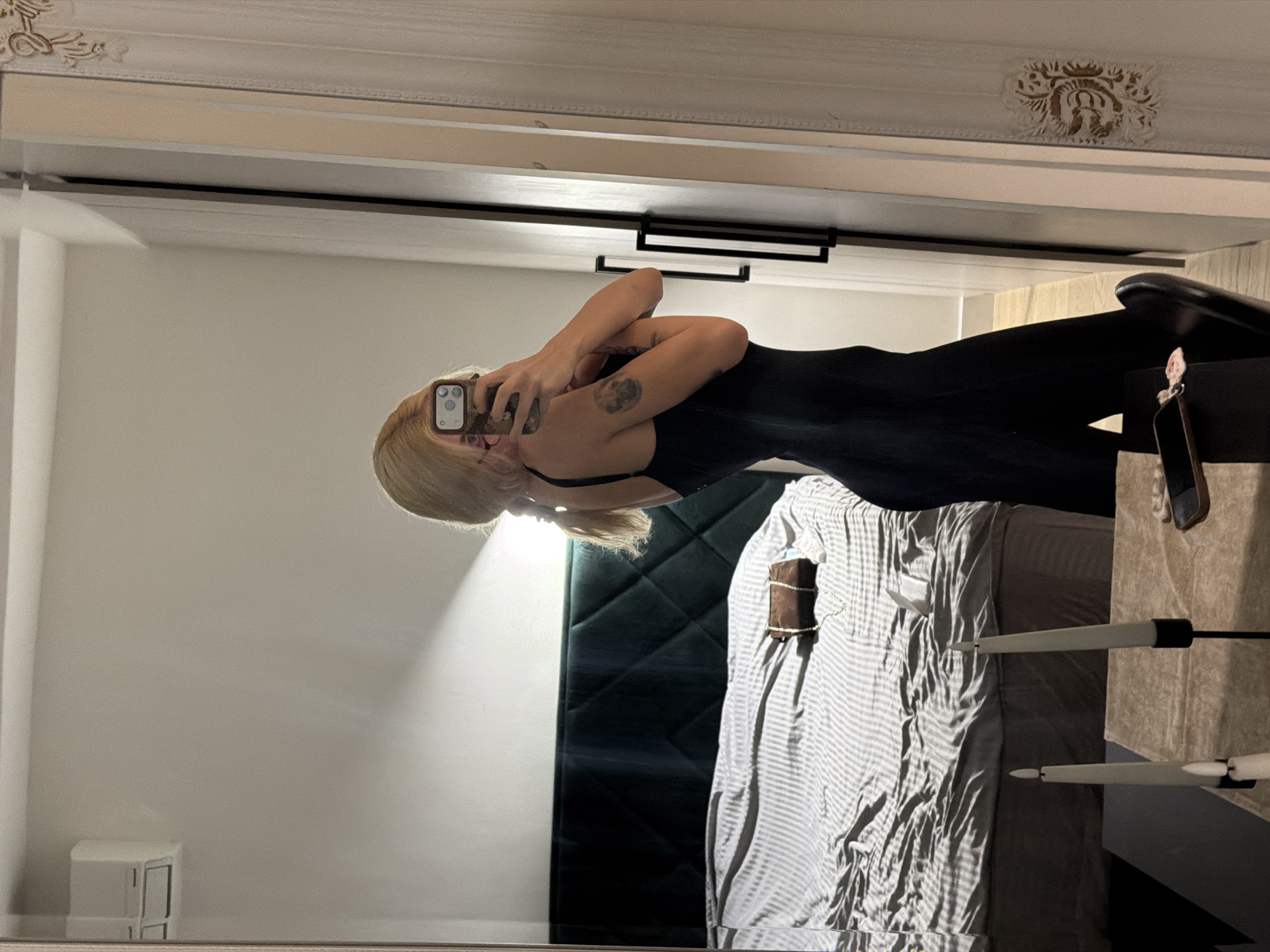 A woman with blond hair is taking a selfie in a mirror. She is wearing a black bodysuit. Her body is angled to the right and she is facing towards the mirror with her phone in her hands. Her hands are near her face. A bed is visible behind her with striped sheets and a dark headboard. A bedside table is also visible. The location is likely a bedroom in a residence. There is no visual information available to determine the specific location in Belgrade, Serbia.