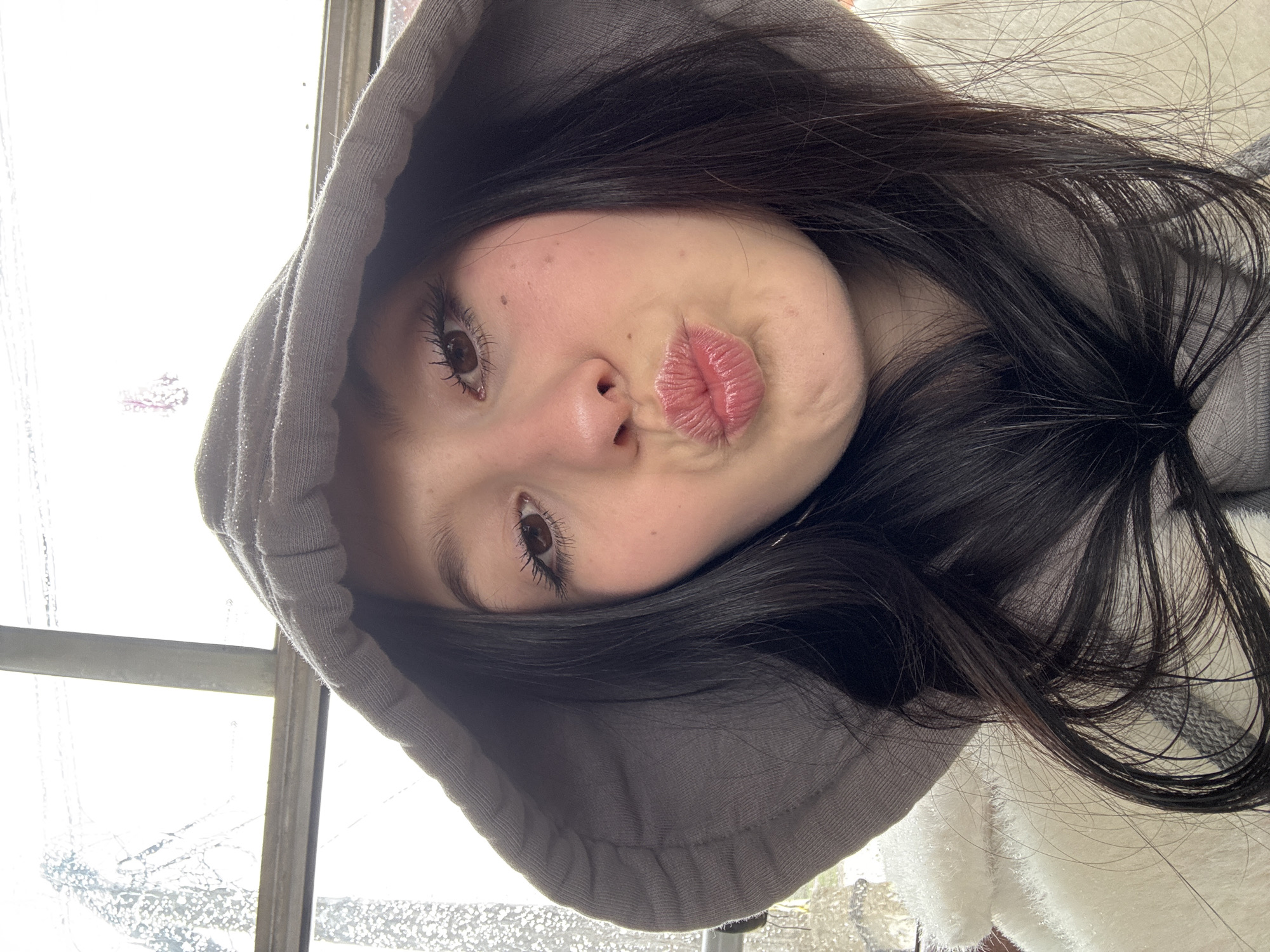 A person with dark hair and a grey hooded sweatshirt is making a kissing face. The individual is facing towards the lower right corner of the frame. A window is visible in the background, suggesting an indoor setting. The location of Фоки, Russia cannot be determined from this image.