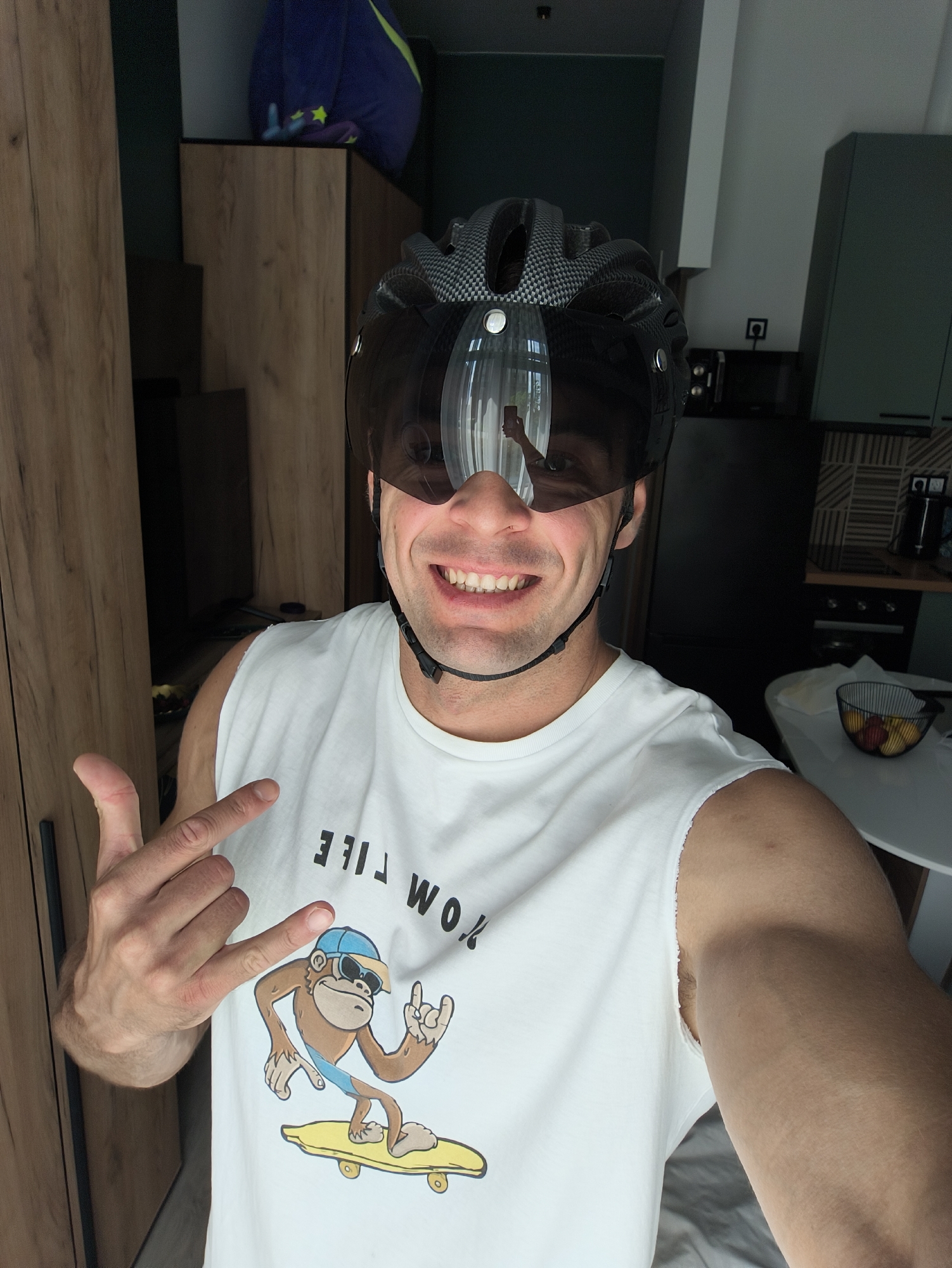 A man is posing for a selfie, wearing a black and grey cycling helmet with a dark visor. He's wearing a white tank top with a cartoon of a monkey skateboarding and the words "WORK LIFE" printed on it. He is giving a "rock on" hand gesture with his left hand. The background shows an indoor scene with furniture including wooden cabinets and a table with a fruit bowl. The location cannot be definitively determined as Moscow, Russia, based on the visual information provided. There is no identifiable landmark or cultural artifact that confirms the specific city or country.