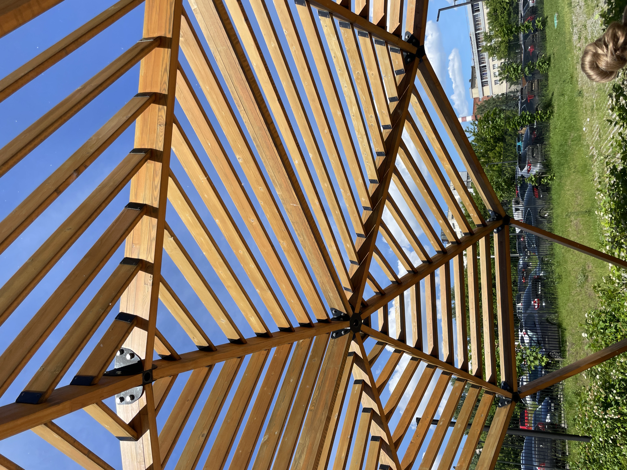 A wide-angle, low-perspective photograph captures the underside of a large, hexagonal or octagonal wooden pergola or pavilion structure against a bright blue sky. The structure is constructed from numerous parallel wooden slats of light brown hue, supported by thicker radiating wooden beams. Black metal plates and L-shaped brackets secured with silver bolts connect the wooden members, notably at a central hub overhead and at points where the slats meet the main beams. The central connection point shows a circular black metal plate with multiple visible bolts.
Through the gaps in the wooden structure, the sky is predominantly clear blue with scattered small white cumulus clouds. Below the structure, in the foreground, lush green grass and several bushes with small white blossoms are visible. In the bottom right corner, the top of a human head with light brown hair styled in a bun is partially visible.
Beyond the immediate foreground, a black metal fence separates the grassy area from a parking lot filled with numerous parked vehicles, primarily dark-colored sedans and SUVs. Several young trees are planted along the fence line. In the midground and background, various multi-story urban buildings are visible. A distinctive tall, slender industrial chimney with alternating horizontal red and white stripes stands prominently against the sky. Other buildings exhibit typical urban architecture. The scene depicts a sunny daytime environment in the city of Izhevsk, Russia.