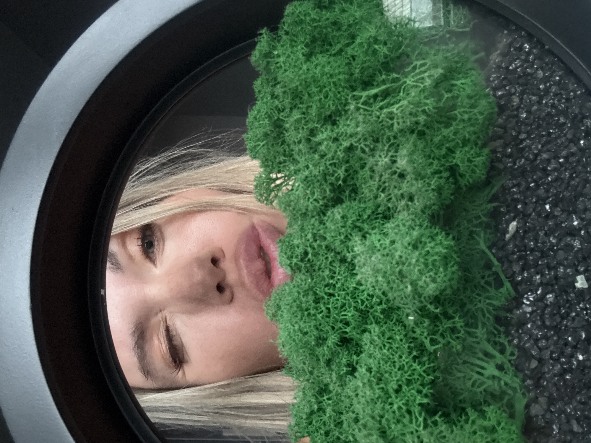 A close-up, vertically oriented image captures a woman's face partially obscured by vibrant green moss within a circular frame. The woman, with light blonde hair, looks toward the left side of the frame. Her left eye is open and alert, while her right eye is partially closed or squinted. Her lips are slightly parted, conveying a subtle expression that could be interpreted as playful or curious.
The green material dominating the right side of the frame is textured, resembling preserved or artificial reindeer moss. To the immediate right of the moss, filling the remainder of the circular frame's interior, is a mass of dark, granular material, consistent with small decorative stones or gravel. The circular frame itself features a distinct dark inner edge and a wider, lighter gray outer band.
The arrangement suggests a decorative display, possibly a terrarium, a unique mirror, or an art installation, creating an intimate and somewhat surreal portrait. There is no specific activity depicted beyond the woman's presence and gaze into the frame. The time of day and weather conditions are not discernible. No text is visible in the image. The image was taken in Votkinsk, Russia.