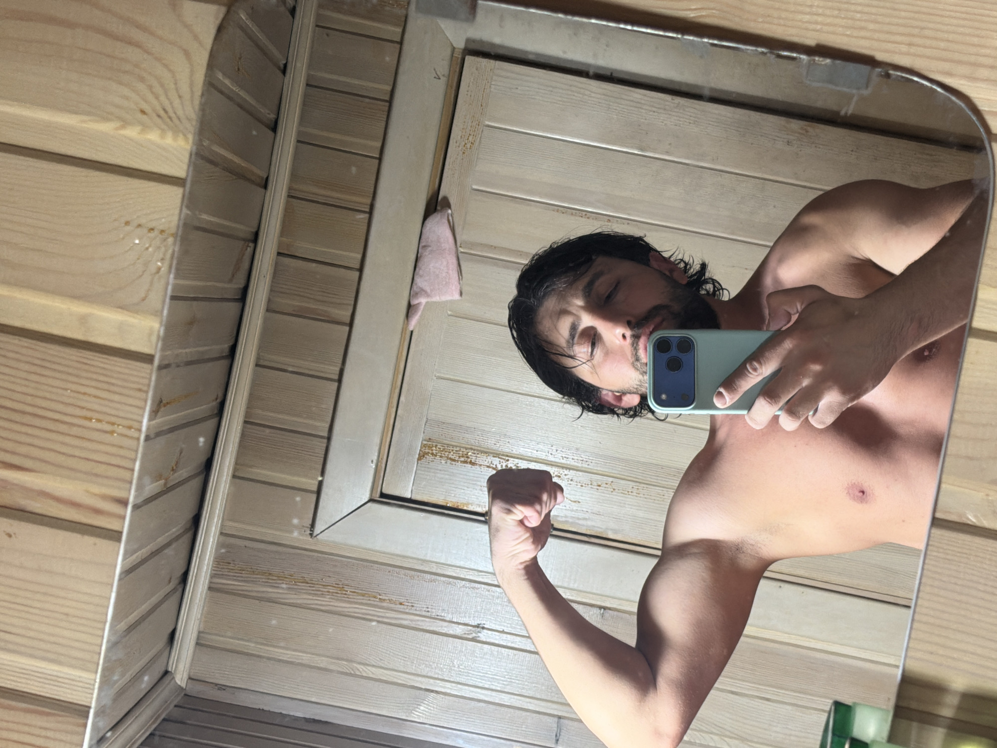 A bare-chested man is posing in front of a mirror while holding a smartphone. The reflection shows his face and flexed arm. He appears to be in a room with wood paneling. The location in the city of Тартат, Russia, is not discernible from the image.