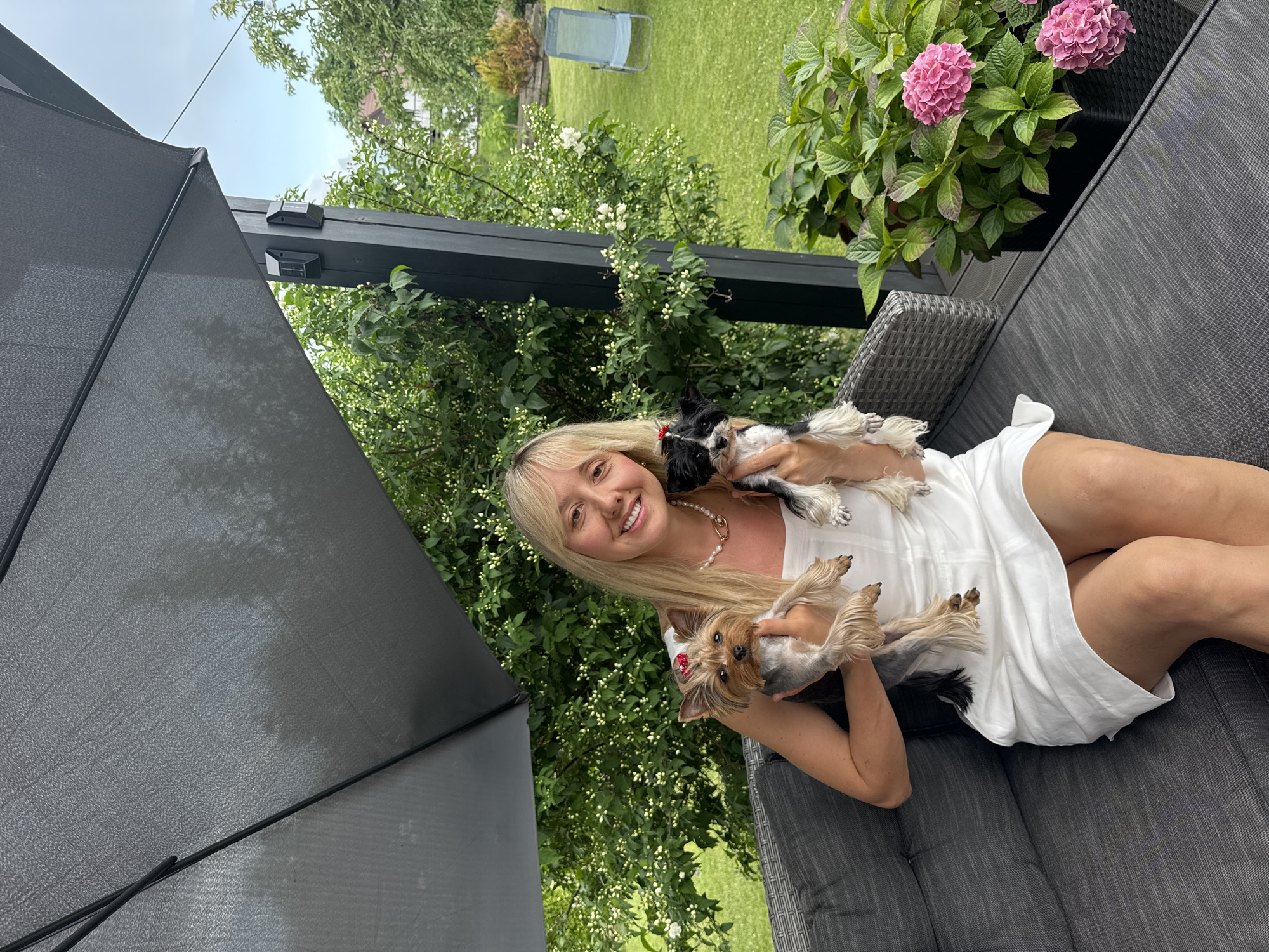 In Carnikava, Latvia, a young blonde woman is seated on an outdoor sofa, holding two small dogs. One dog, a Yorkshire Terrier, rests on her lap, looking towards the camera with a red bow on its head. Another small dog, black and white, is nestled against her shoulder, its head resting on her arm. The woman is wearing a white dress and a pearl necklace. She is smiling, appearing relaxed and happy.
The setting is a backyard garden on a cloudy day. Lush green trees and shrubs surround the area. In the background, a lawn is visible, with a blue lounge chair placed on it. A pot of pink hydrangeas sits on the deck beside the sofa, adding a splash of color to the scene. Part of a dark grey umbrella is visible on the left side of the frame, suggesting it may be providing shade. The overall atmosphere is peaceful and domestic, conveying a sense of comfort and enjoyment. There is no visible text in the image.