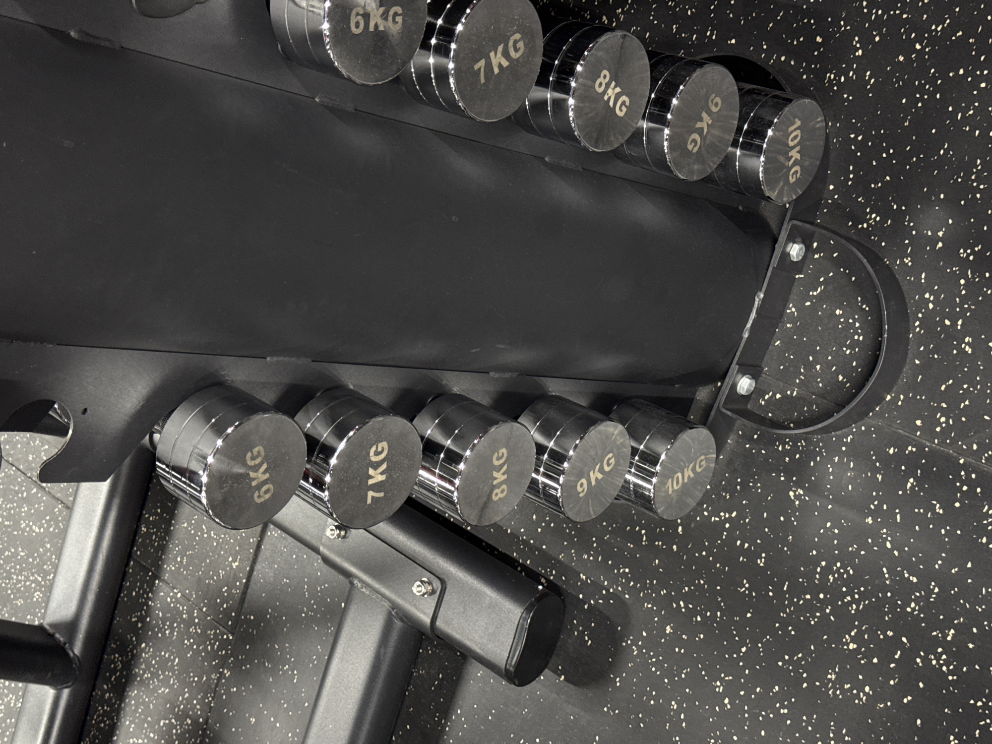 This media file captures a close-up, high-angle view of a dumbbell rack in a gym, likely located in Yerevan, Armenia. The rack, made of dark, matte metal, holds several pairs of dumbbells arranged in ascending order of weight. The dumbbells themselves are chrome-plated with black rubber grips and feature the weight clearly marked in kilograms. Visible weights include 6KG, 7KG, 8KG, 9KG, and 10KG.
The dumbbells are lined up neatly, suggesting organization and readiness for use. The gym floor is a dark rubberized material speckled with white and light grey flecks, a common surface in fitness environments for shock absorption and grip. The lighting appears to be artificial, typical of an indoor gym, casting subtle shadows that highlight the reflective surfaces of the dumbbells.
No people are visible in the frame, indicating the image was taken during a quiet period or before the gym opened. The overall mood is one of functionality and order, emphasizing the equipment. The visual focus is on the weights and the rack, showcasing the variety of weights available for workouts. The metallic sheen of the dumbbells contrasts with the matte texture of the rack and the speckled floor. The scene is static and professional, typical of fitness equipment photography.