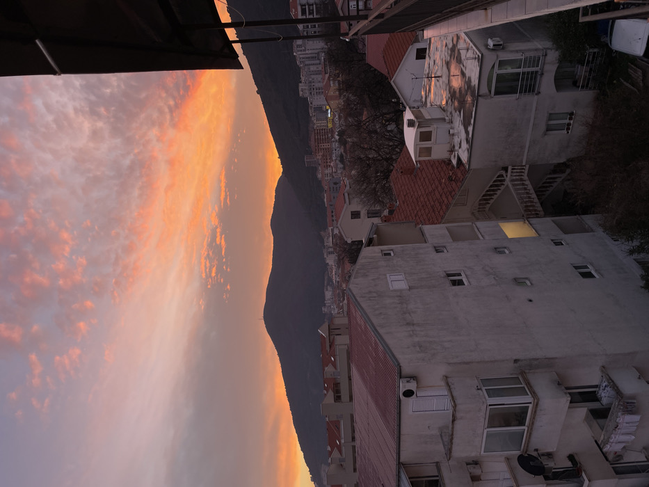 This image depicts a panoramic sunset view over a residential area in Budva, Montenegro. The sky, dominating the upper portion of the frame, exhibits vibrant hues of orange, pink, and purple, with scattered stratocumulus clouds brightly illuminated by the setting sun. Dark, silhouetted mountains form the horizon line, with a visible communication tower atop the peak of the leftmost mountain.
The cityscape below consists of densely packed residential buildings with predominantly red or terracotta-tiled roofs. In the immediate foreground on the left, a weathered, light-colored multi-story building features multiple windows, balconies, external air conditioning units, and a satellite dish. Clothes are visible hanging to dry on a lower-level balcony. A single window on the building's facade emits a distinct yellow light. To the right, lower-rise structures with red-tiled roofs are interspersed with one building featuring a flat roof that reflects the sunset colors, suggesting a wet surface. An external concrete staircase with white railings descends between buildings in the mid-ground. Sparse, bare deciduous trees are visible among the structures. The perspective is from an elevated position, likely a balcony, indicated by a dark overhead structure and railing partially visible in the upper right. No individuals or explicit interactions are discernible within the scene.