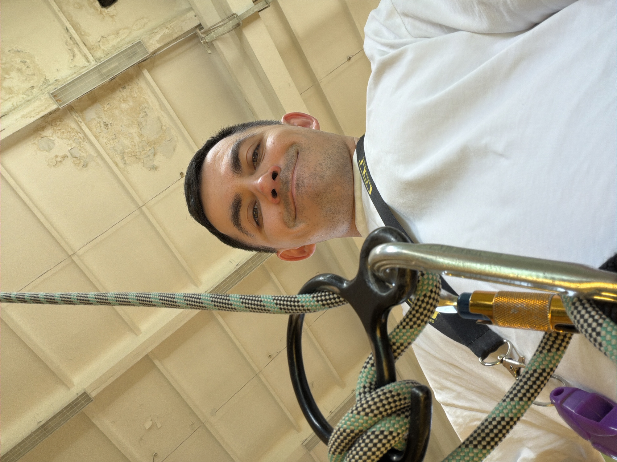 A low-angle indoor shot looking up at a man wearing a white t-shirt and a harness. He has dark hair and a slight smile. The harness is connected to climbing gear, including rope, a black belay device, a gold-colored locking carabiner, and a purple plastic object. The background consists of a paneled ceiling with water damage and some overhead lighting. The image does not provide information on the location in the city of Izhevsk, Russia.