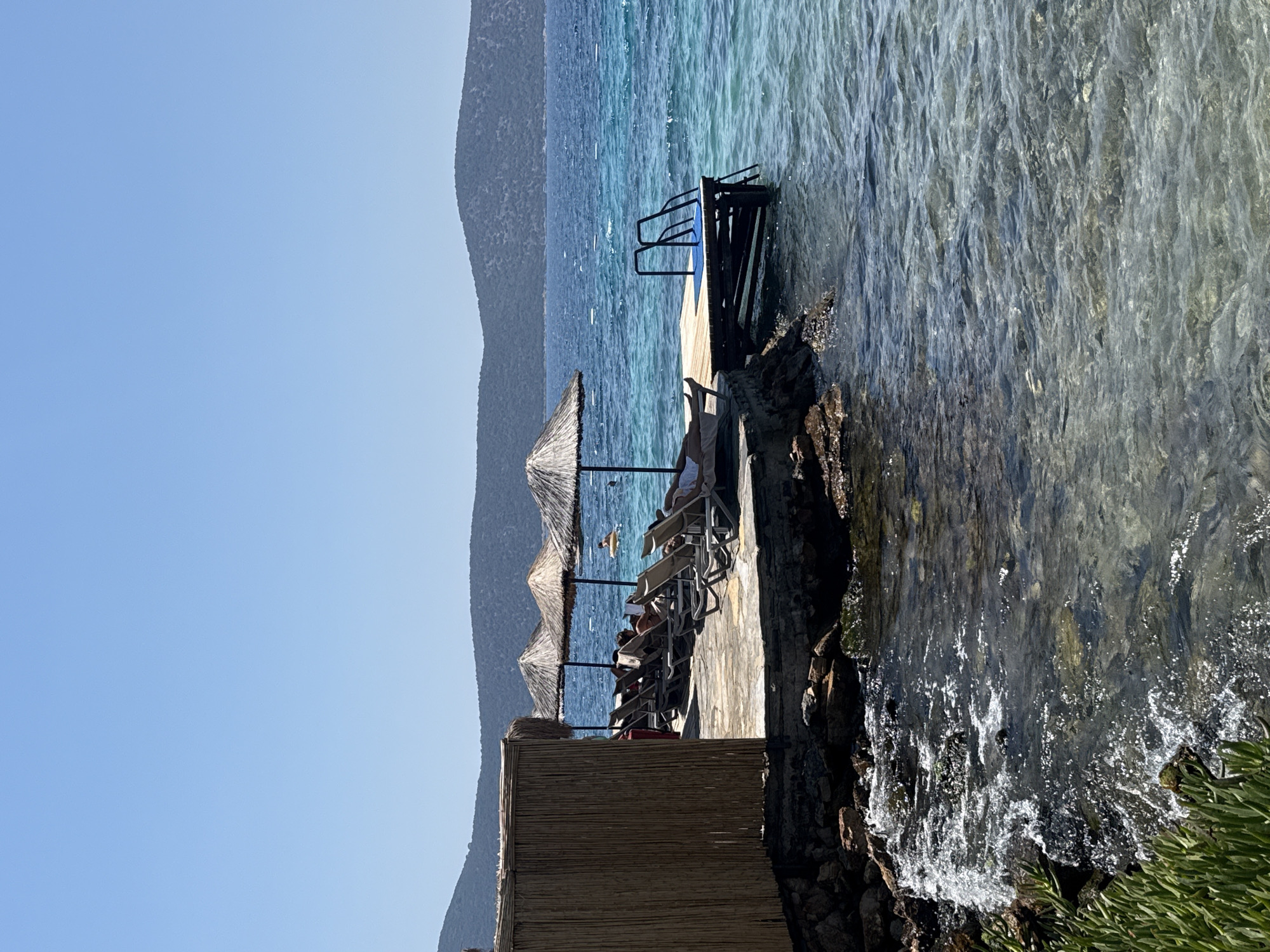 The image depicts a serene waterfront scene in Bodrum, Turkey, on a clear, sunny day. In the foreground, the turquoise water gently laps against a rocky shore lined with lush green vegetation. Further out, a wooden pier extends into the sea, equipped with a ladder for swimmers. Several straw umbrellas provide shade for lounge chairs arranged on a stone platform overlooking the water, where individuals are seen relaxing. In the background, rolling hills covered in greenery form a picturesque backdrop against the vast expanse of the blue sky. The overall ambiance is one of a tranquil, seaside retreat, characteristic of the popular tourist destination.